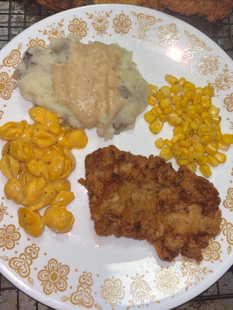 A plate of food with fried chicken, mashed potatoes topped with gravy, yellow corn, and creamy macaroni and cheese on a white floral-patterned plate.