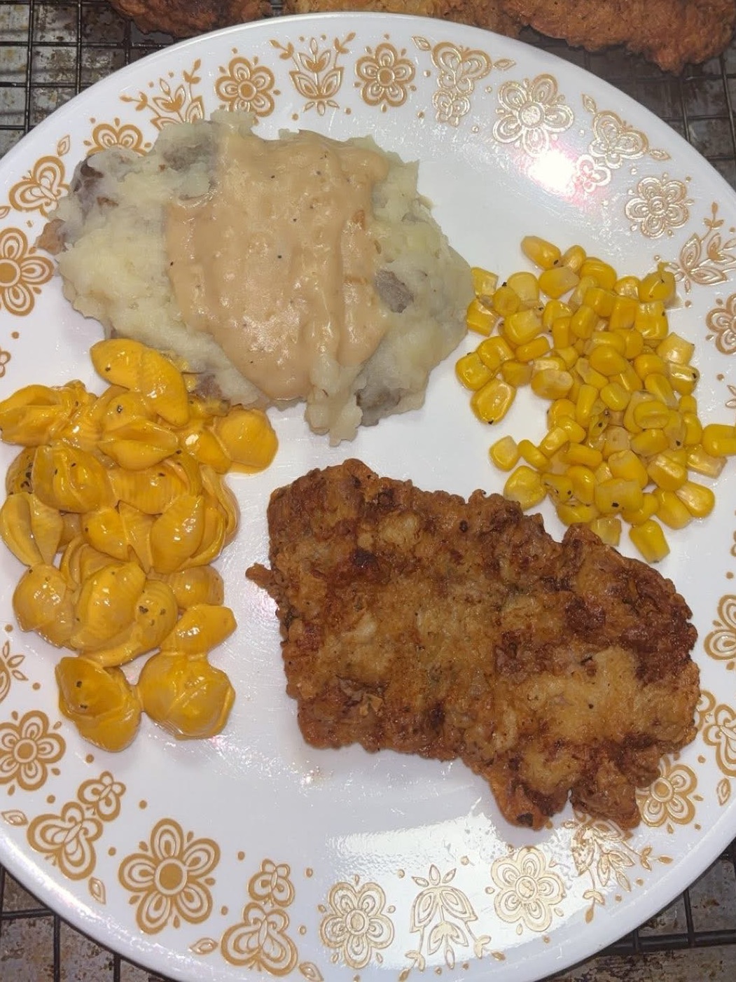 A plate of food with fried chicken, mashed potatoes topped with gravy, yellow corn, and creamy macaroni and cheese on a white floral-patterned plate.