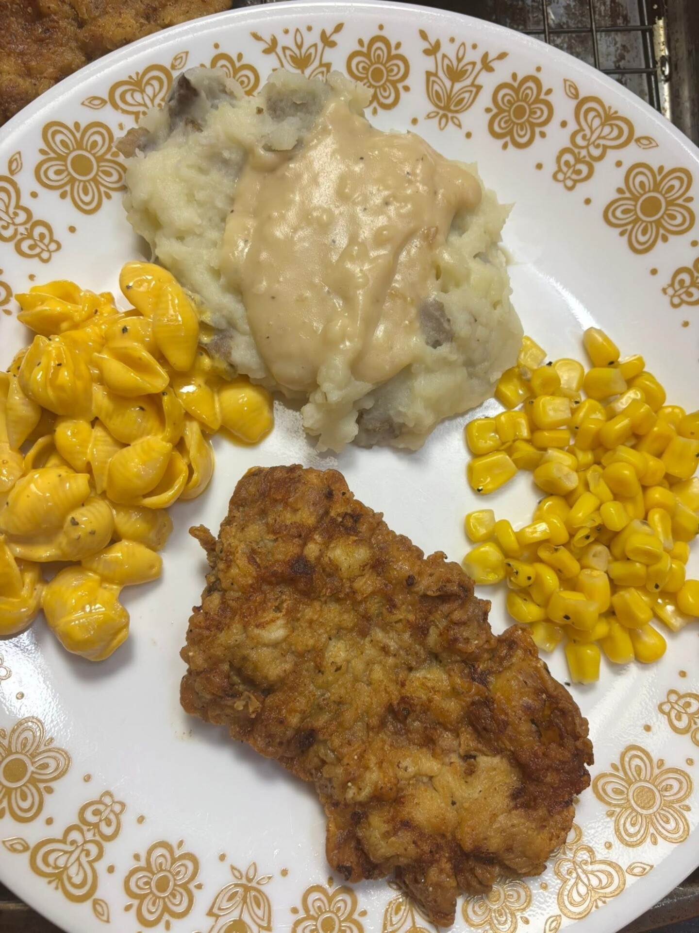 A plate with fried chicken steak, mashed potatoes topped with gravy, creamed macaroni and cheese, and yellow corn. The plate has a brown floral pattern around the edge.