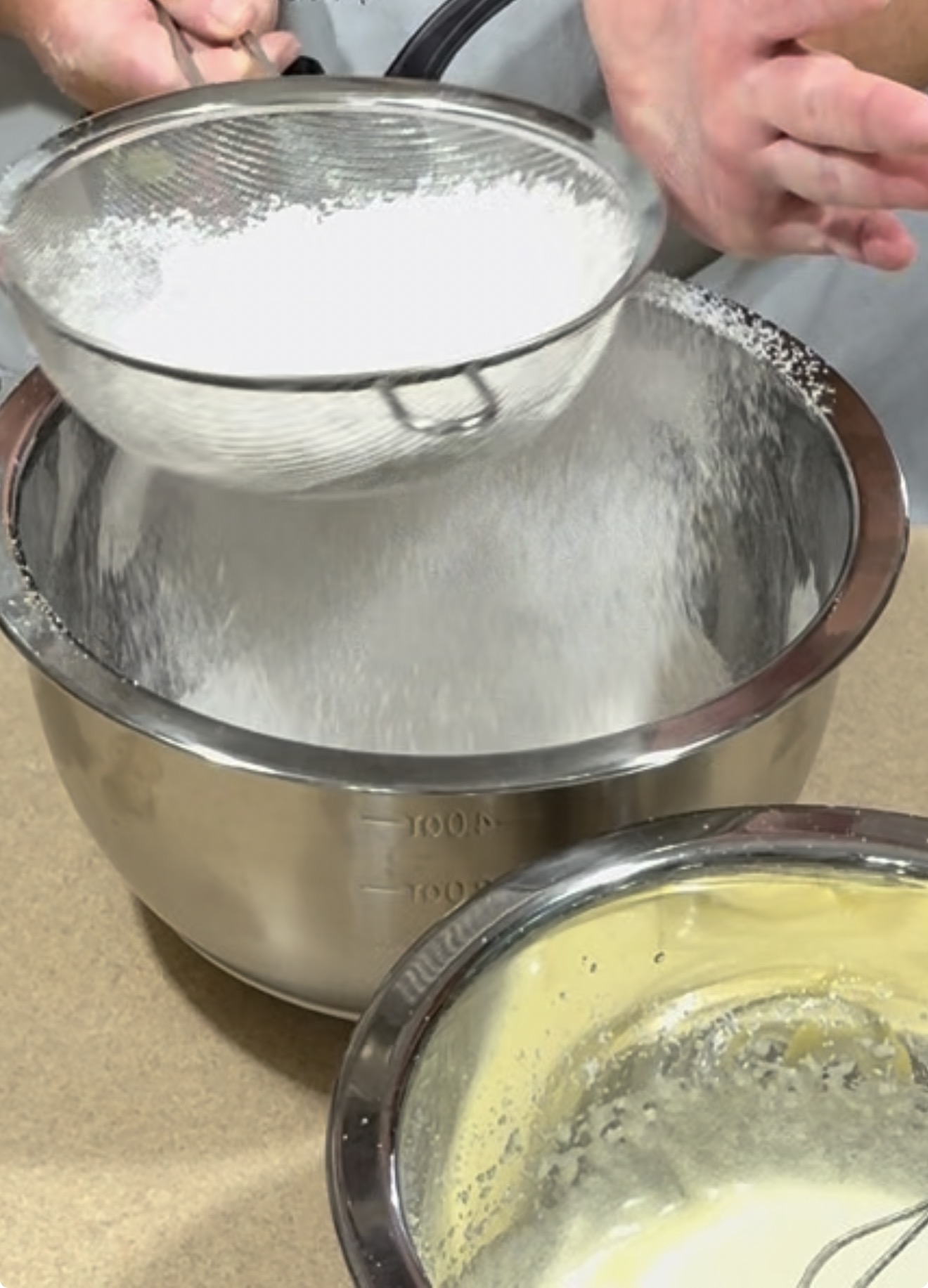 A person sifts flour or powdered sugar into a large metal mixing bowl, with another bowl containing a yellow batter visible on the countertop nearby.