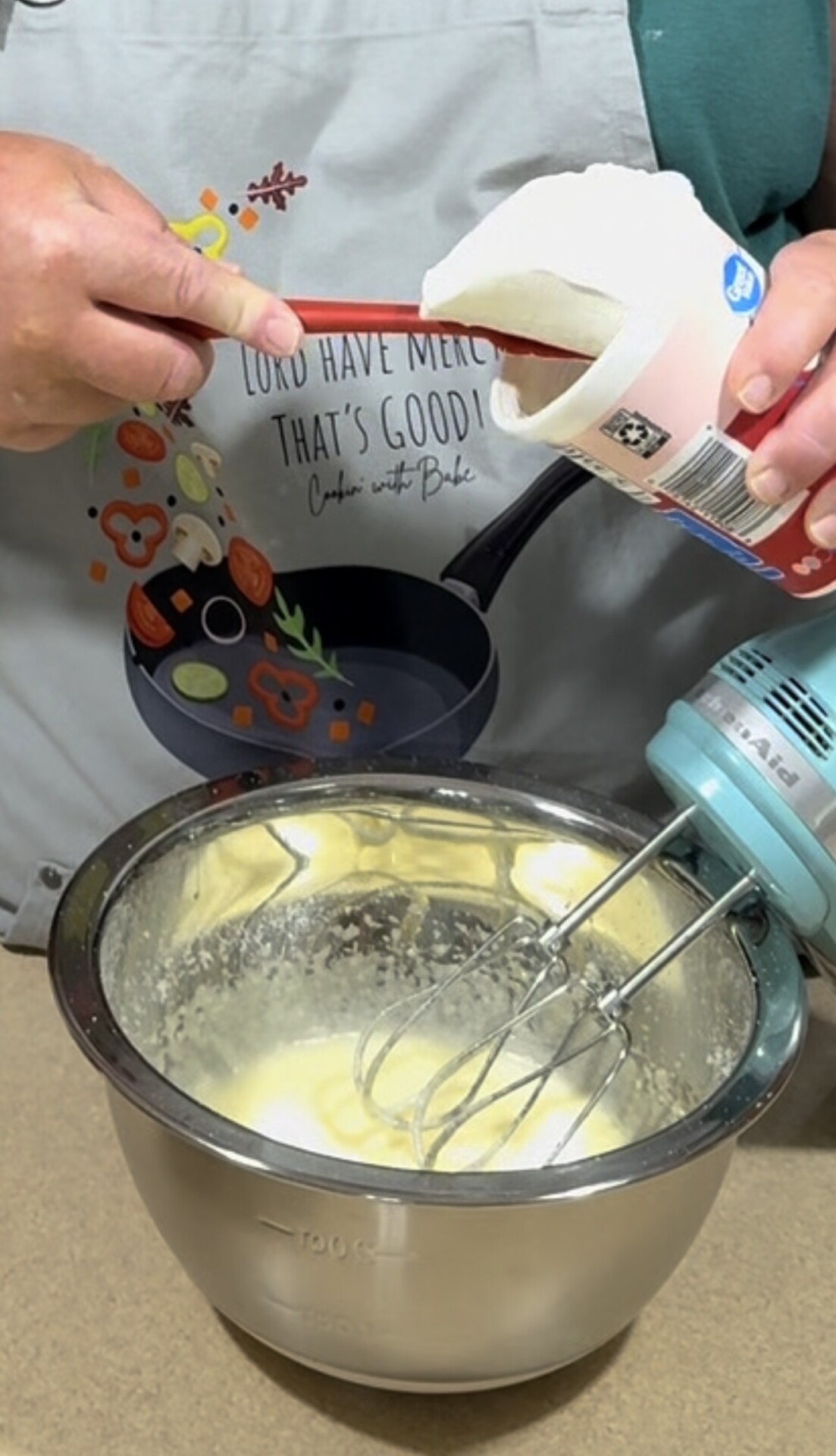 A person in an apron uses a spatula to scoop sour cream from a container into a mixing bowl, with an electric hand mixer resting in the bowl.