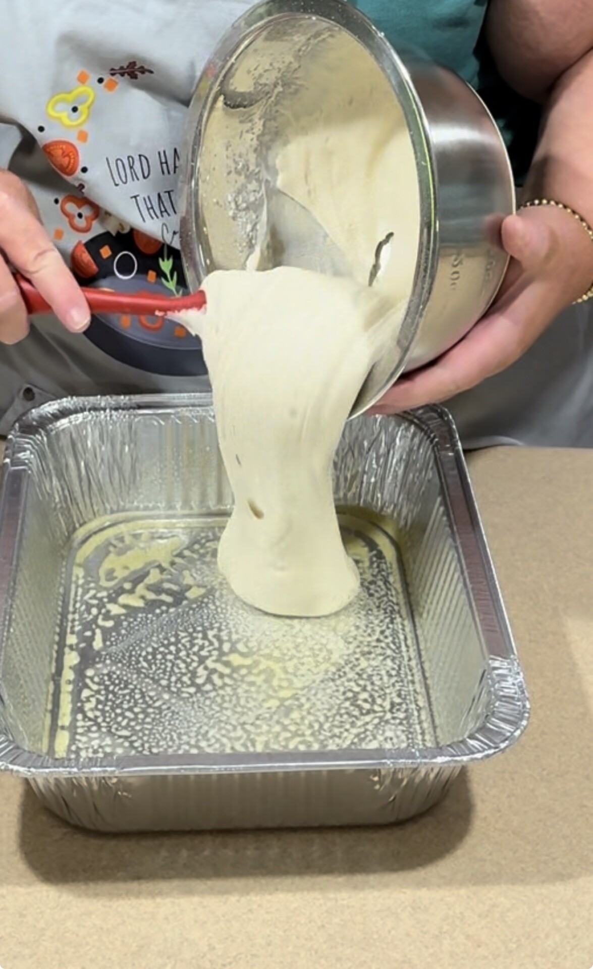 A person pours cake batter from a metal bowl into a greased, square aluminum baking pan on a kitchen counter.
