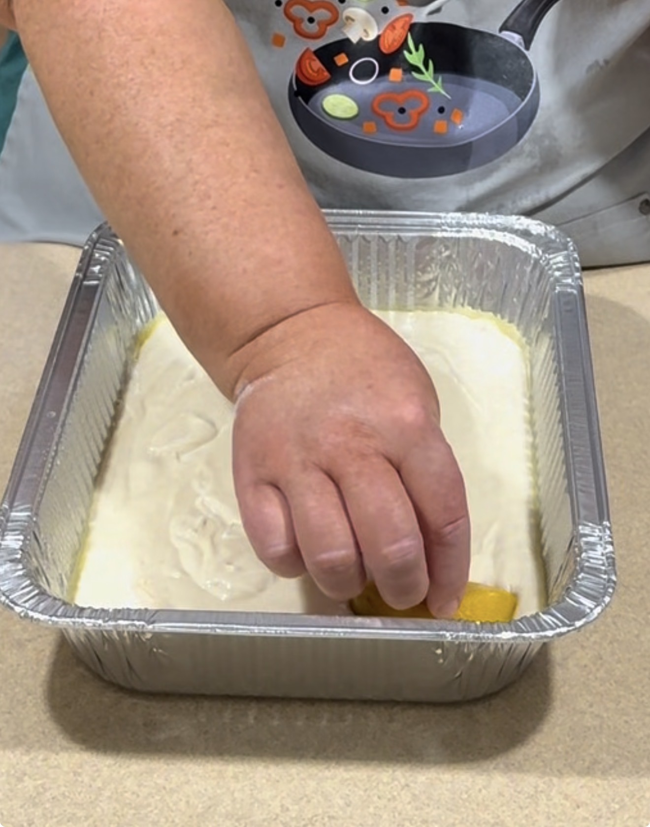 A person places a sliced peach onto a pan filled with cake batter. The pan is aluminum and sits on a countertop, and the person is wearing an apron with a cooking-themed design.