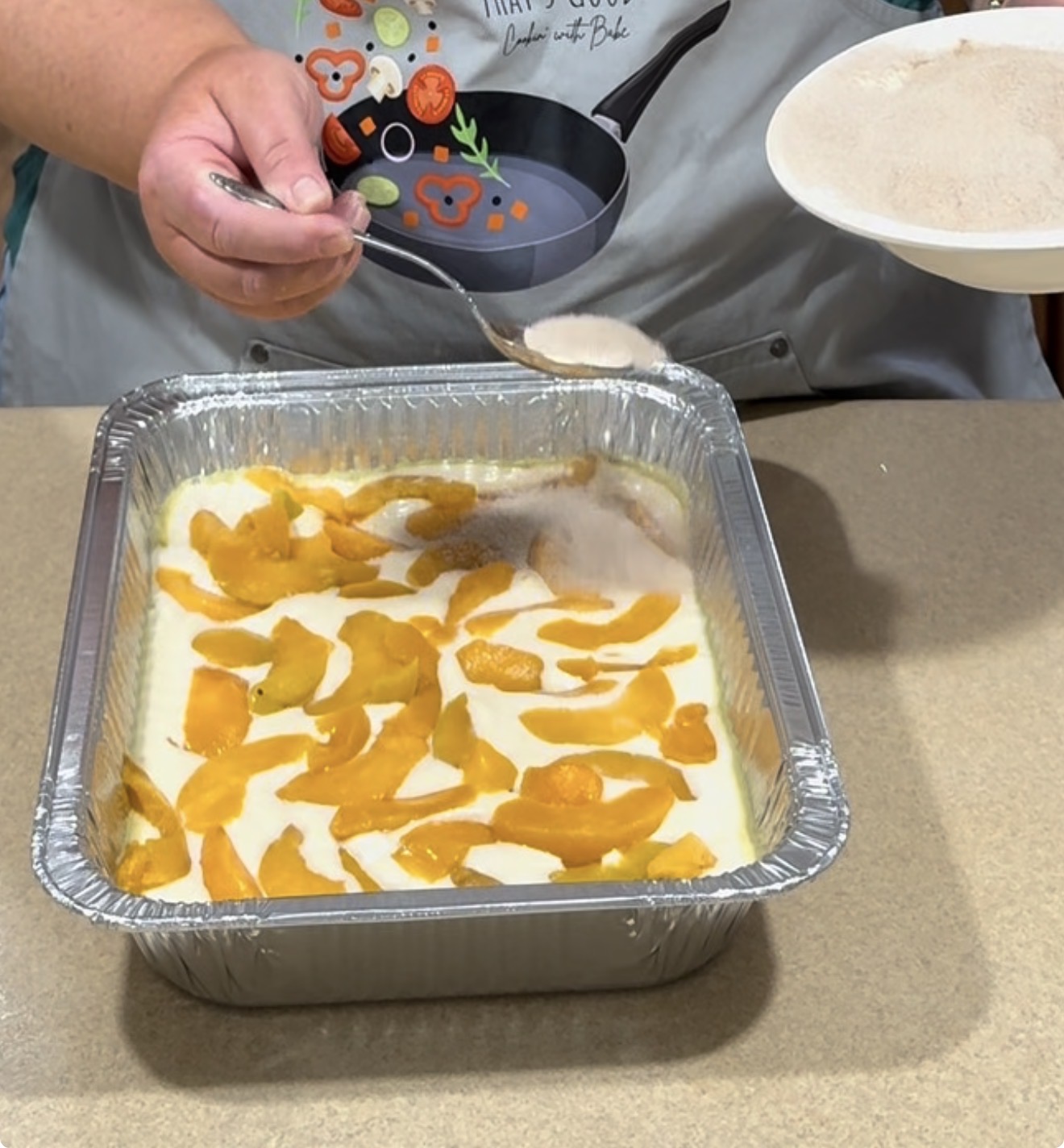 A person sprinkles sugar from a spoon over a baking pan filled with sliced peaches and batter. The pan sits on a countertop, and the person wears an apron with cooking utensils printed on it.