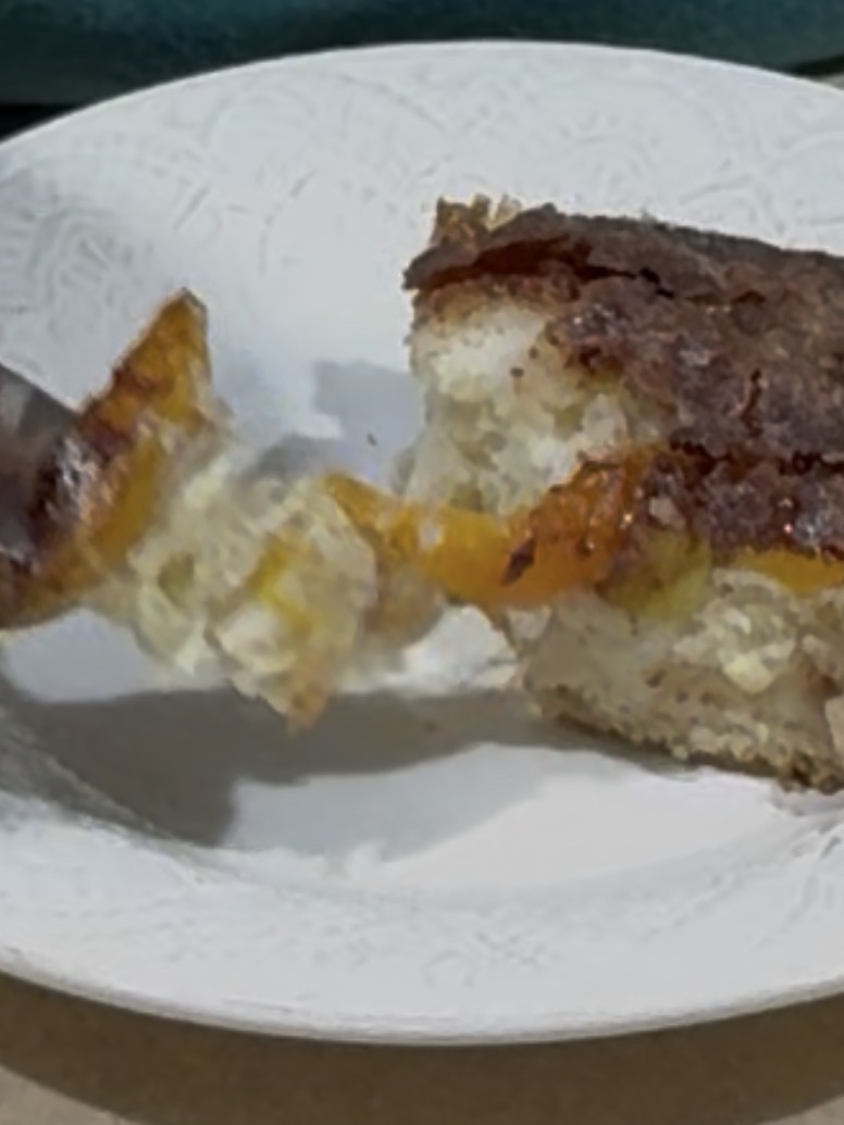 A close-up of a fork taking a bite from a slice of cake with a yellow fruit layer and a brown topping, served on a white patterned plate.