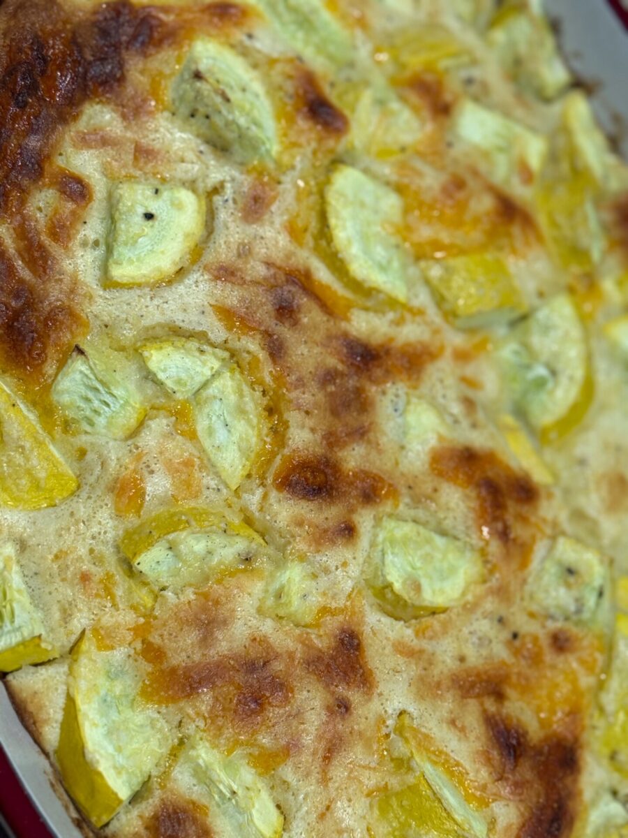Close-up of a baked casserole topped with chunks of yellow squash. The surface is golden brown with bubbly, melted cheese and visible seasoning.