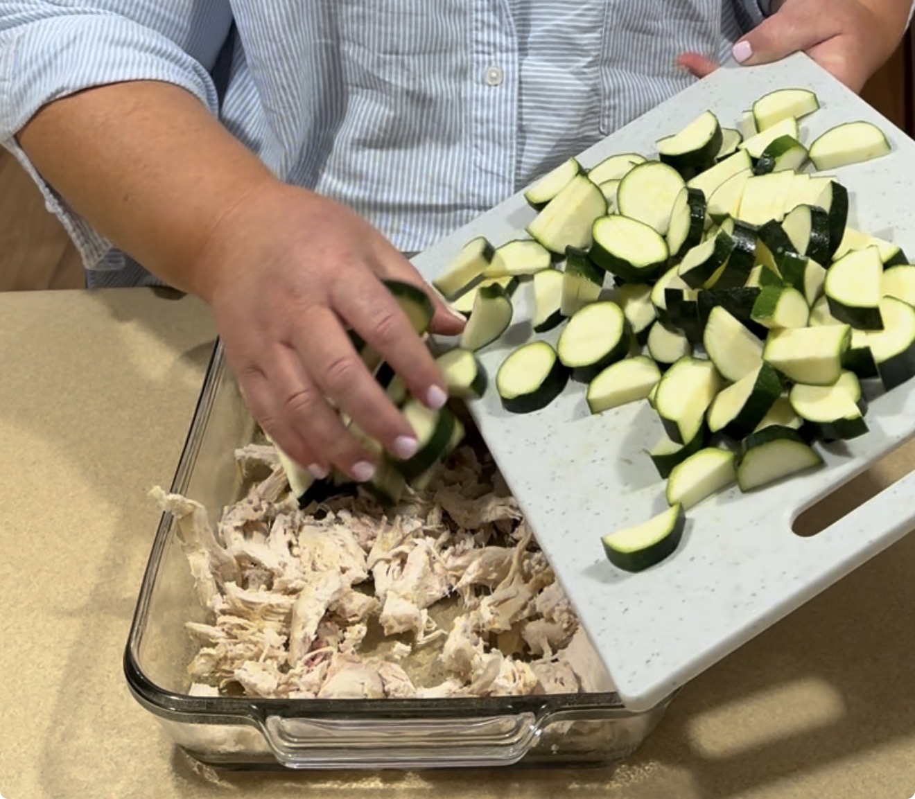 A person pours chopped zucchini from a cutting board into a glass baking dish containing shredded chicken. The person is wearing a light blue shirt with rolled-up sleeves.