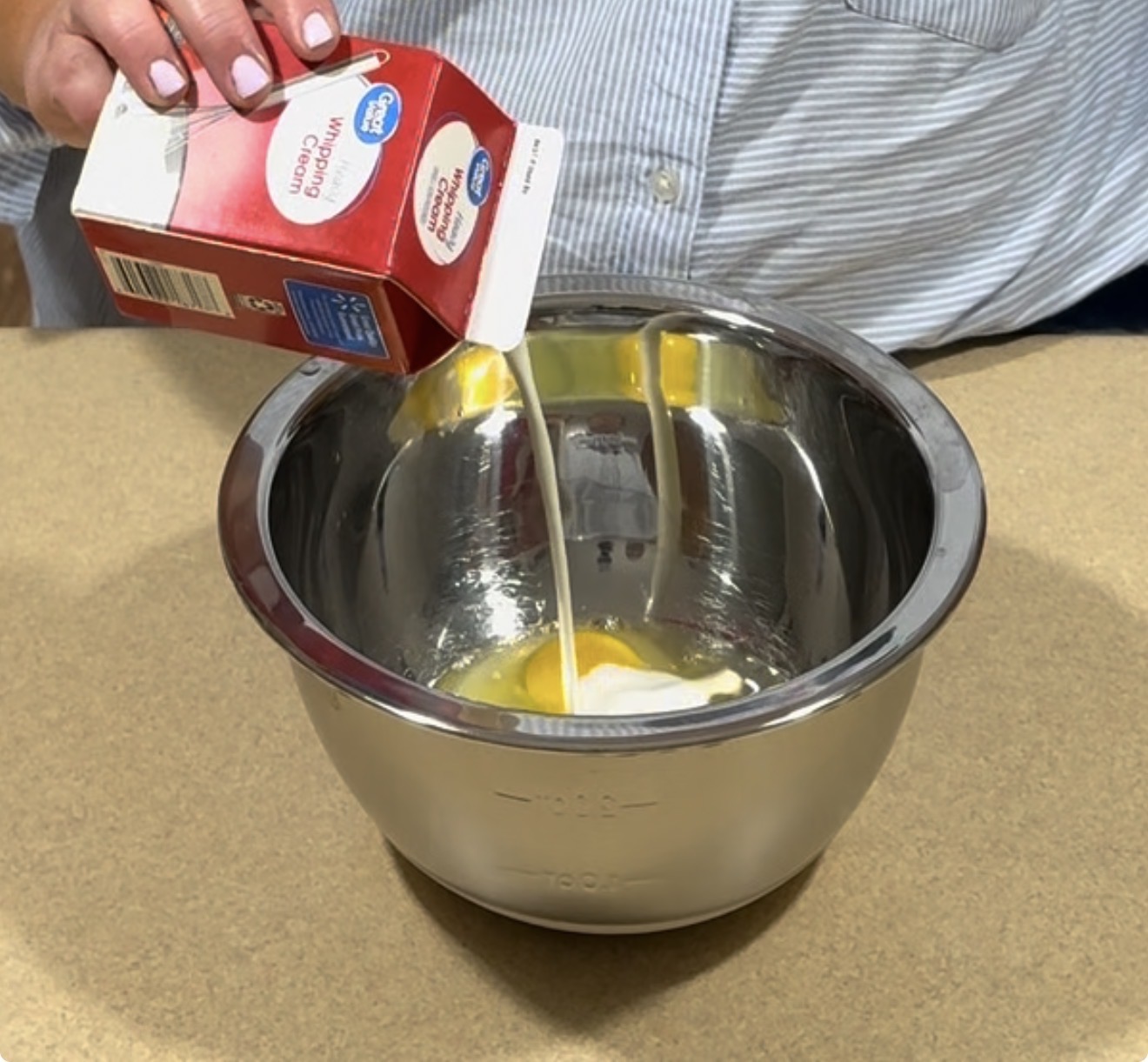 A person pours heavy cream from a carton into a metal mixing bowl containing eggs and sugar on a kitchen counter.