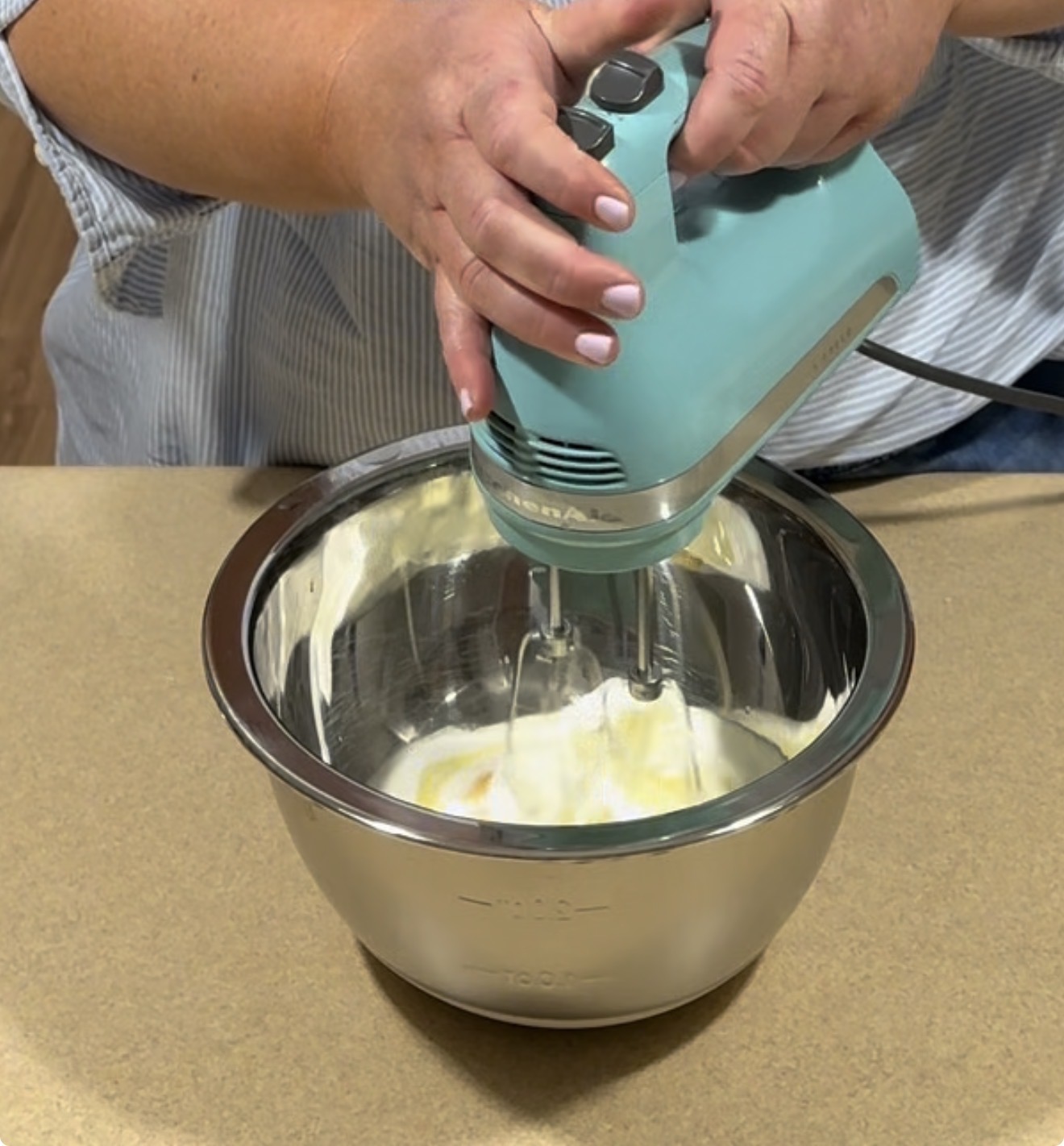 A person uses a teal hand mixer to blend ingredients in a stainless steel mixing bowl on a kitchen counter.