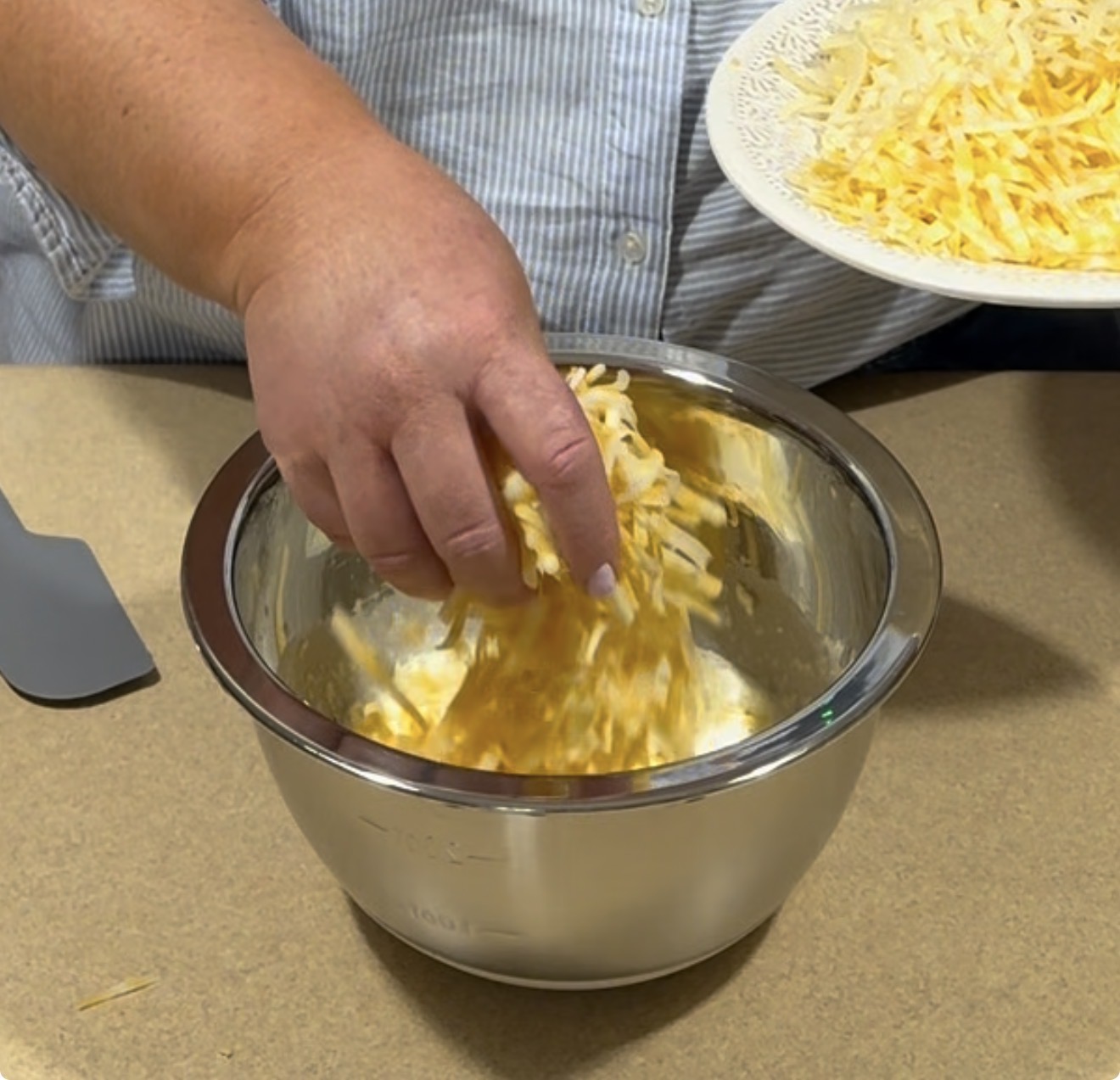A person adds shredded yellow and white cheese from a plate into a stainless steel mixing bowl on a kitchen counter. A spatula rests nearby.