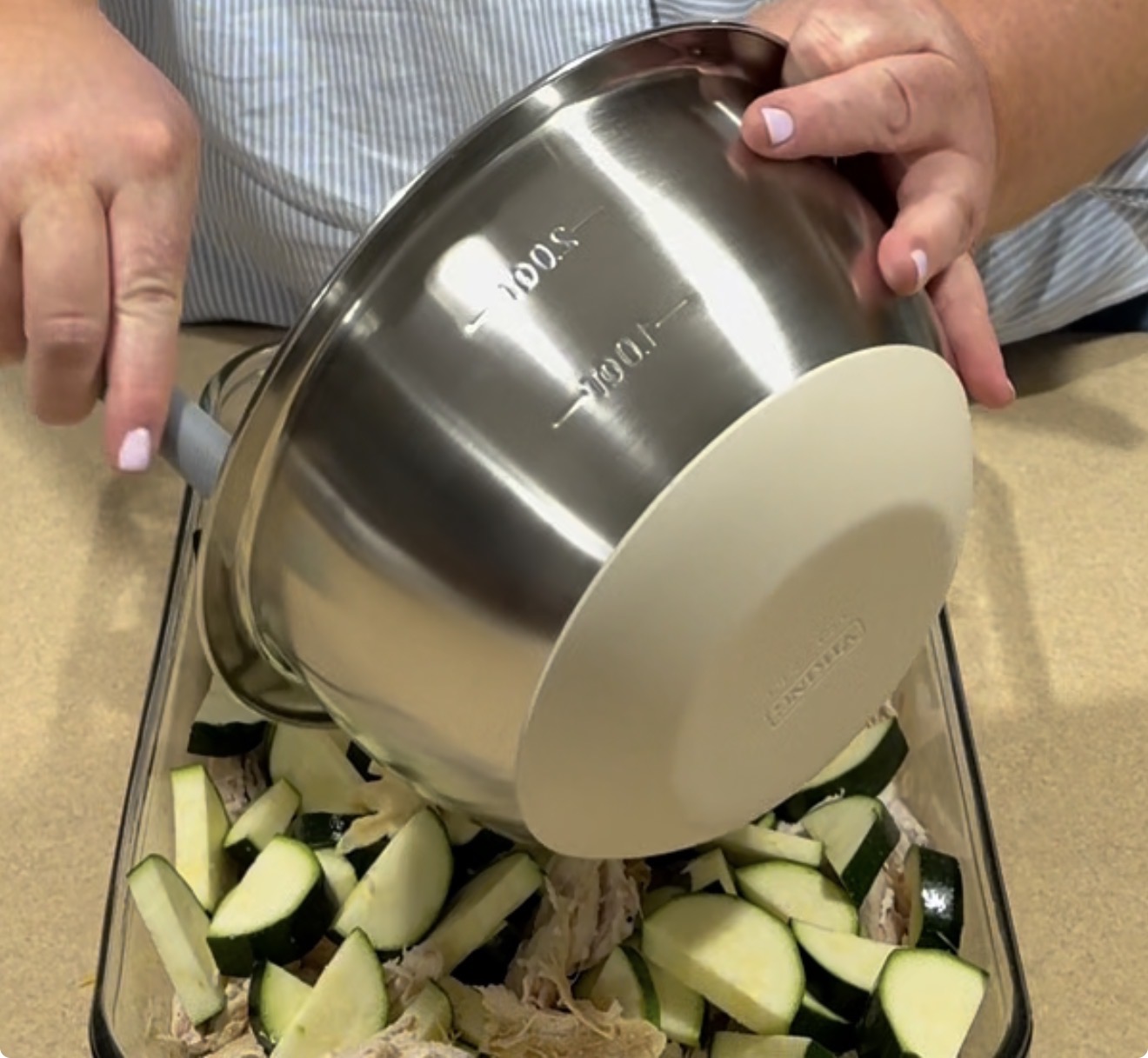 A person pours sliced zucchini from a large stainless steel mixing bowl into a glass baking dish filled with shredded chicken.