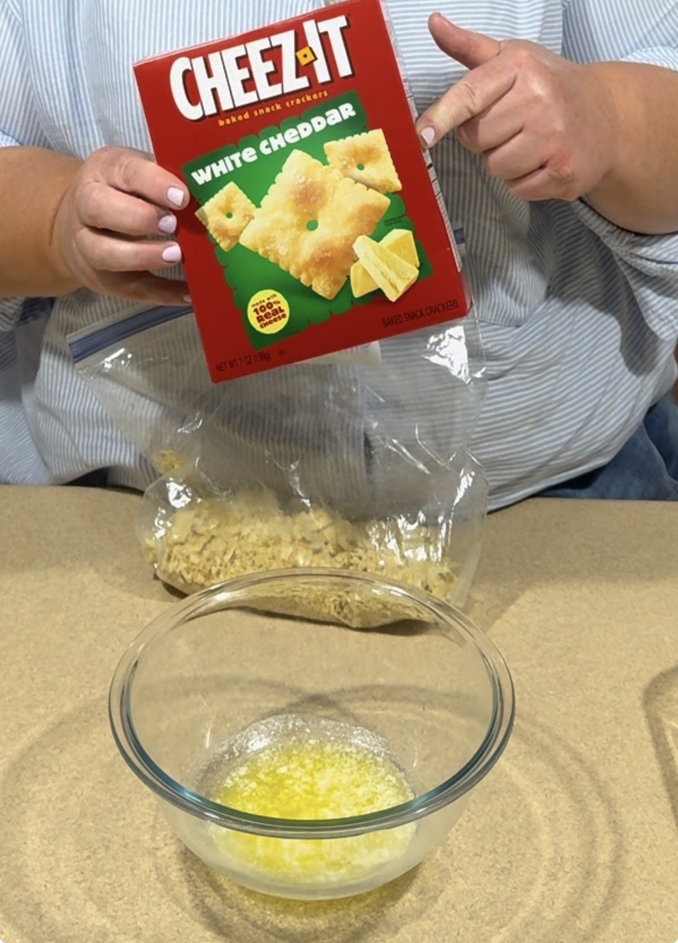 A person holds a box of white cheddar Cheez-It crackers above a plastic bag of crushed crackers, with a bowl of melted butter on the counter in front of them.