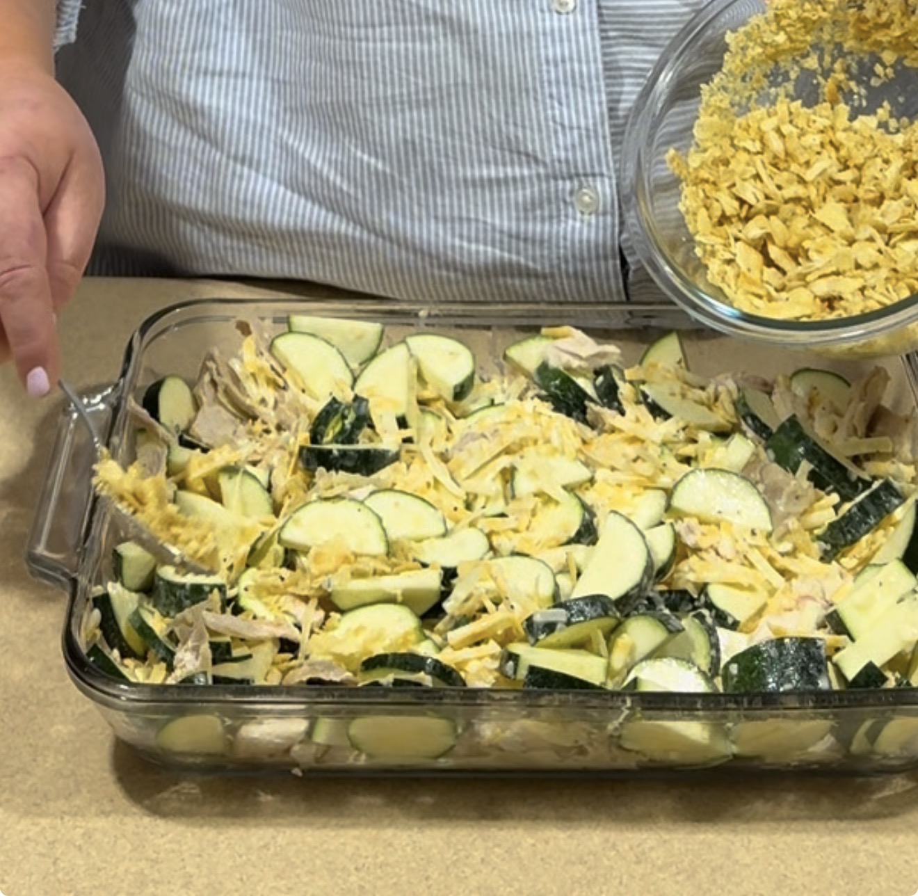 A person sprinkles crushed cornflakes from a bowl onto a casserole dish filled with sliced zucchini, cheese, and shredded chicken.