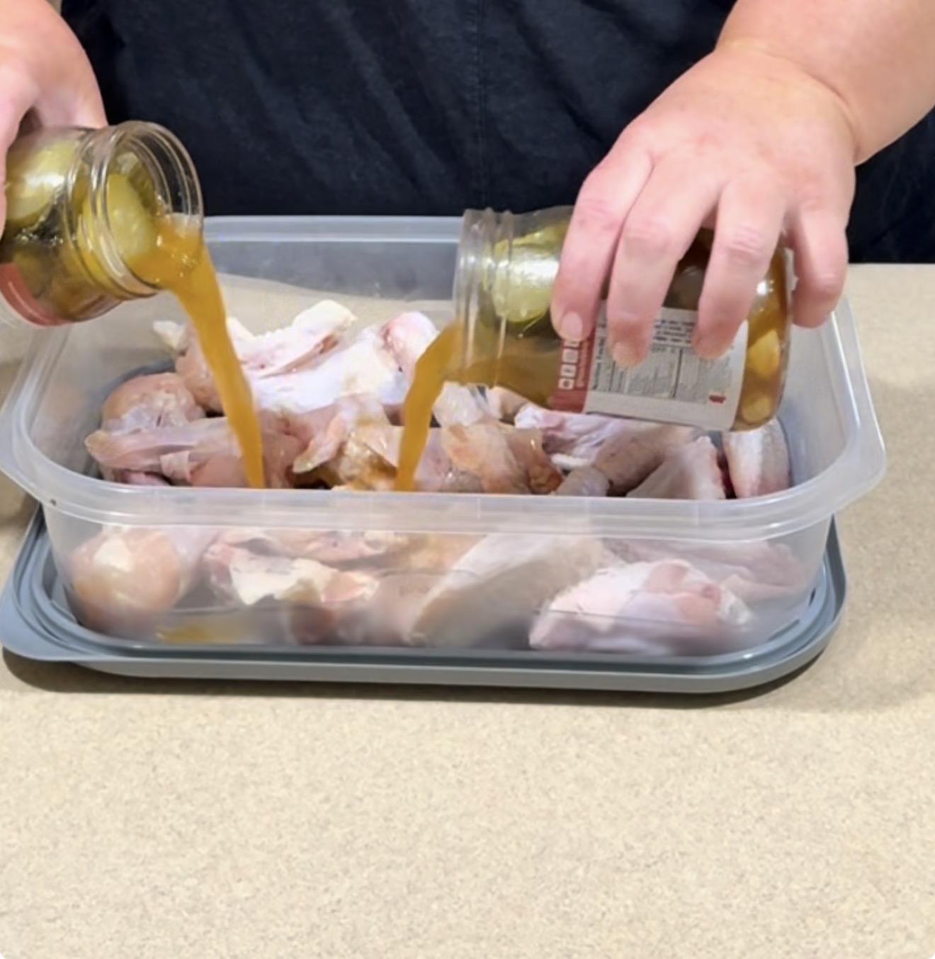 A person pours pickle juice and pickles from a glass jar over raw chicken wings in a plastic container on a kitchen counter.
