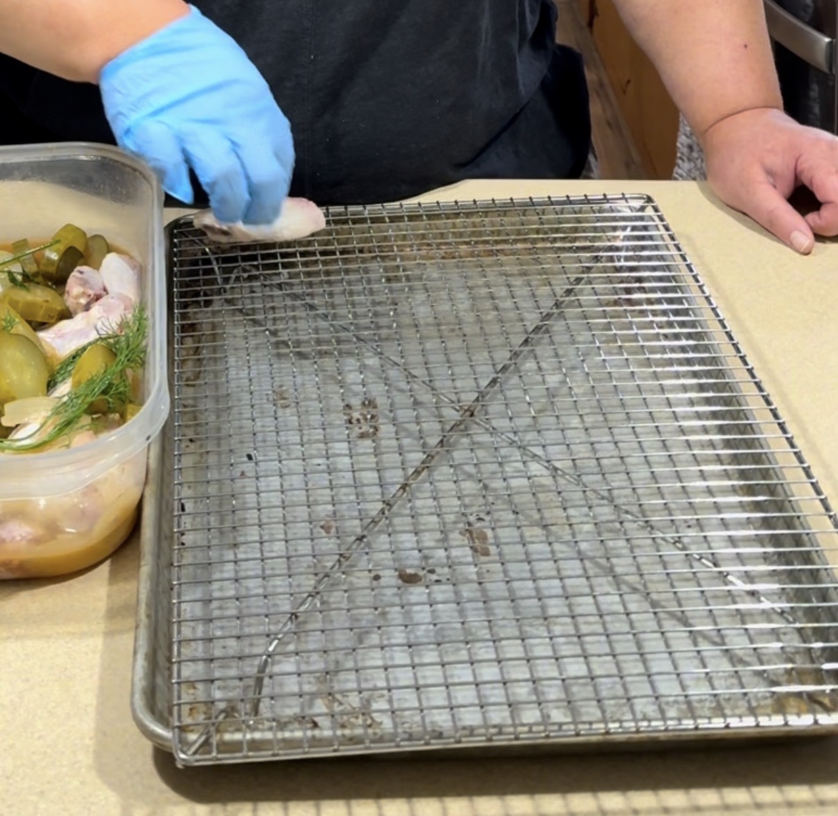 A person wearing a blue glove places marinated chicken from a plastic container onto a wire rack set over a baking sheet on a kitchen counter.