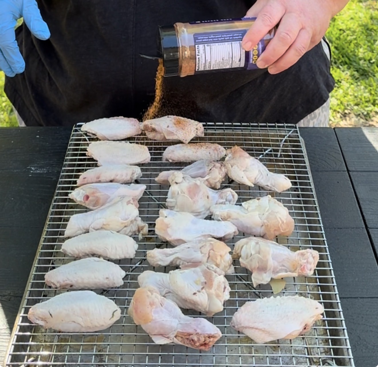 A person sprinkles seasoning from a bottle onto raw chicken wings arranged on a wire rack over a black table outdoors.