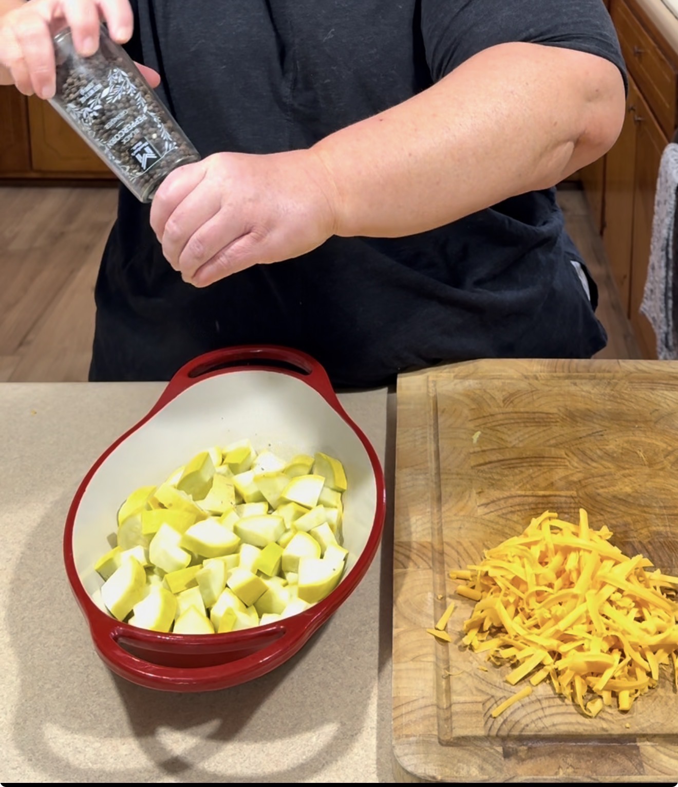 A person seasons chopped yellow squash in a red baking dish, with a pile of shredded cheddar cheese on a cutting board nearby.