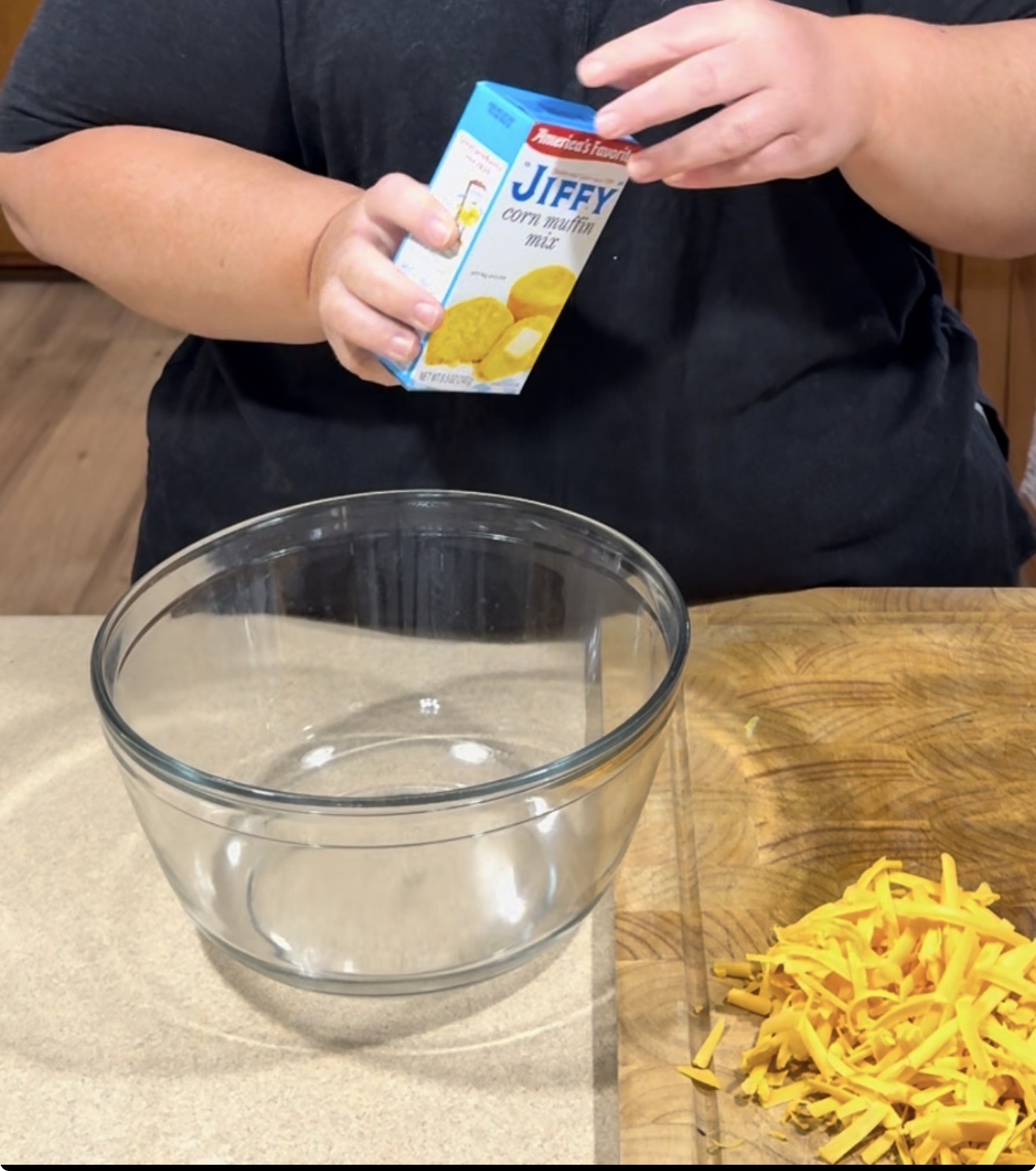 A person holding a box of Jiffy corn muffin mix over a clear glass mixing bowl, with shredded cheddar cheese on a cutting board nearby.
