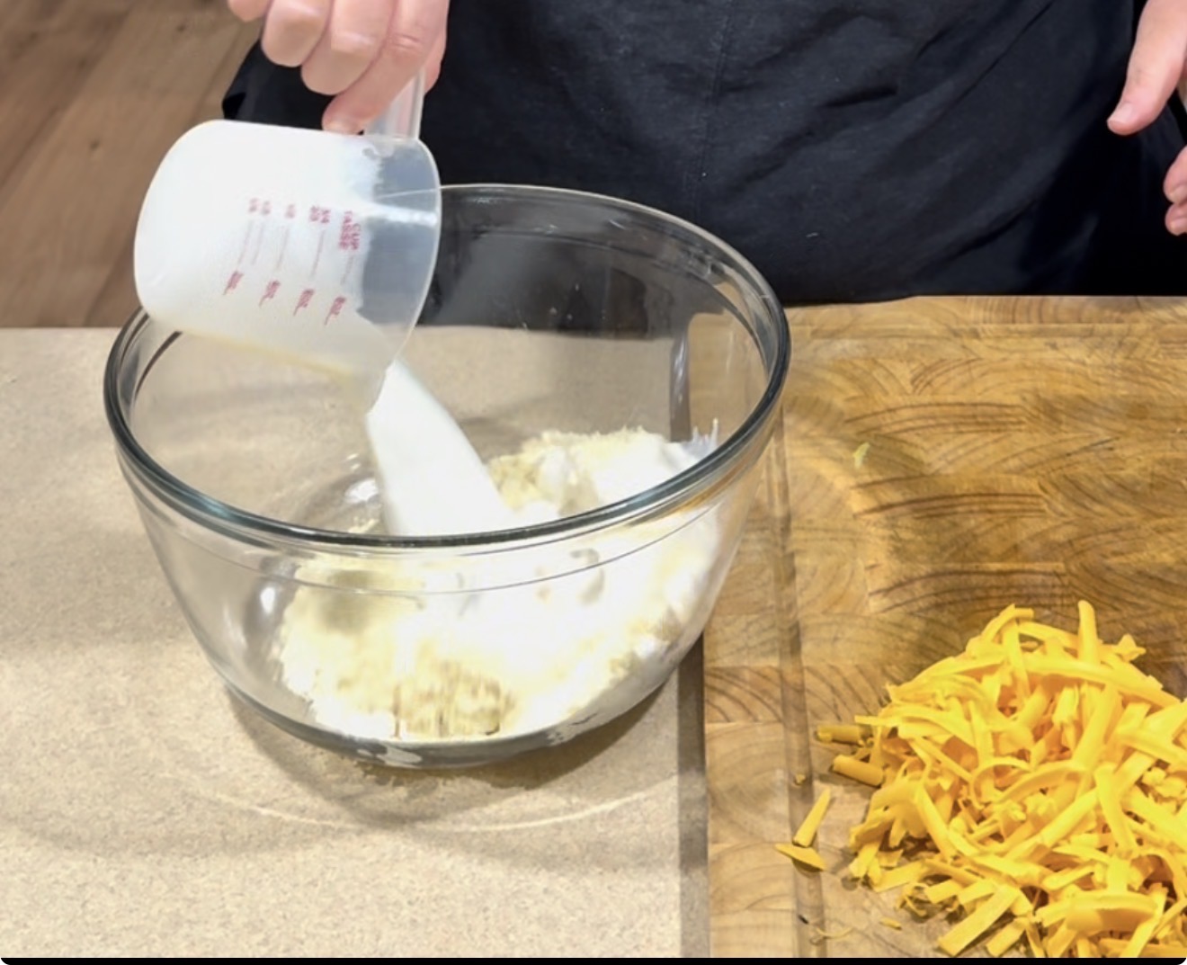 A person pours milk from a measuring cup into a glass bowl with flour mixture on a counter. Shredded cheddar cheese is on a wooden cutting board beside the bowl.