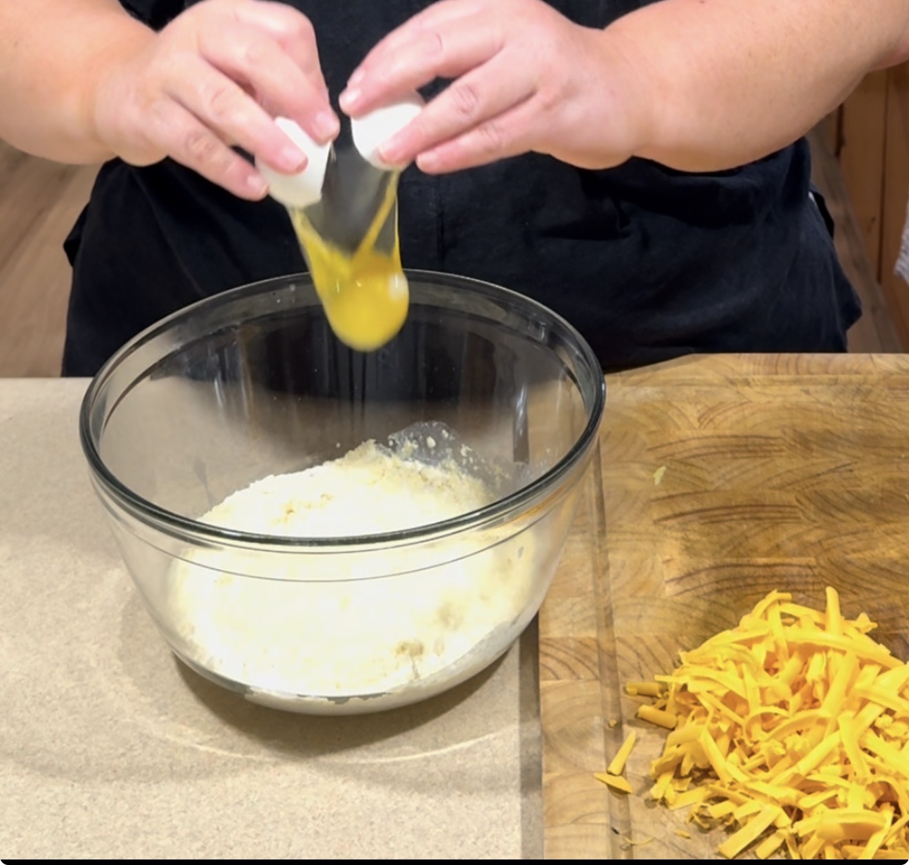 A person cracks an egg into a glass mixing bowl filled with dry ingredients, with shredded cheddar cheese on a wooden cutting board nearby.