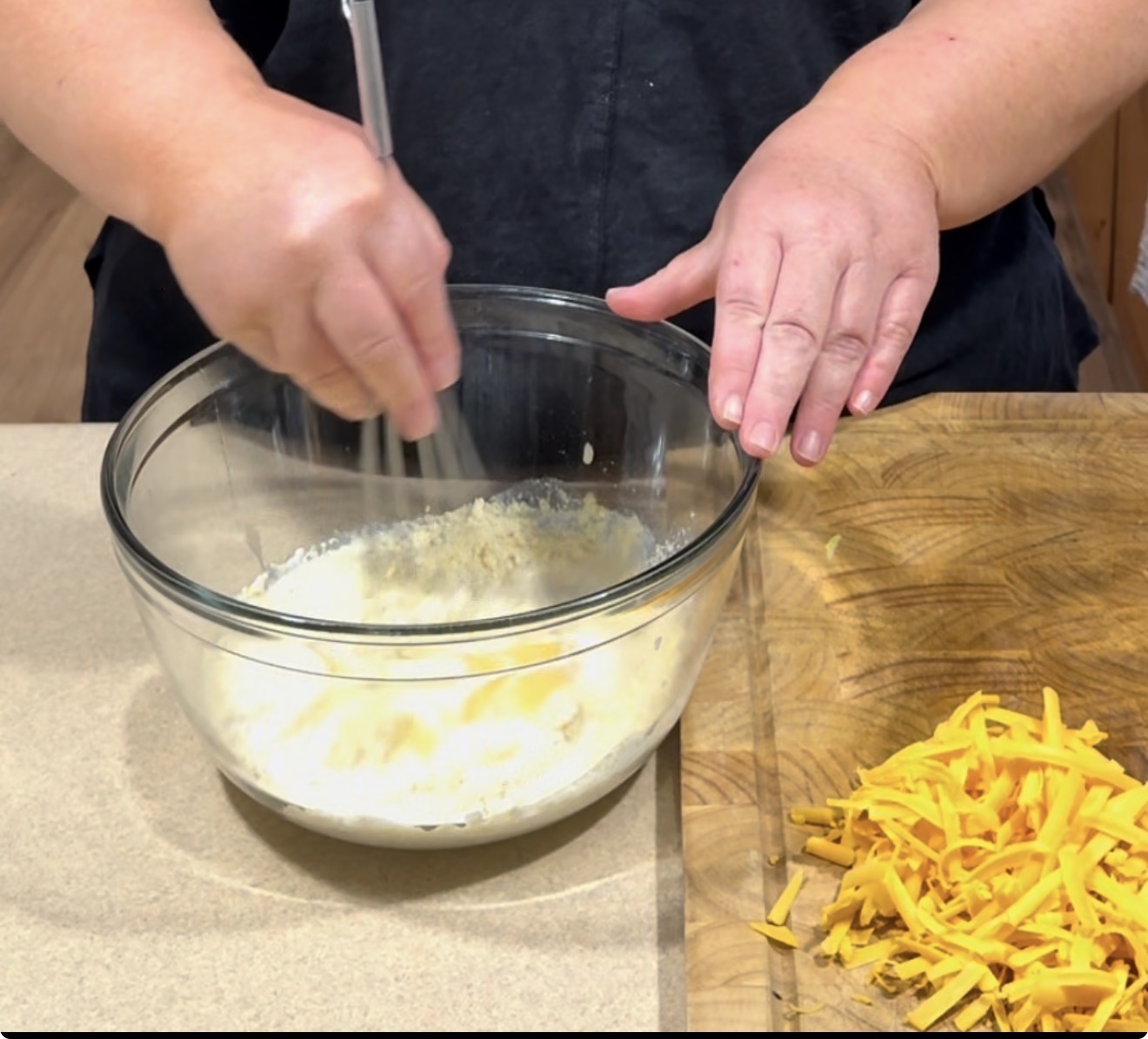 A person whisking ingredients in a glass bowl on a countertop, with a pile of shredded cheddar cheese on a wooden cutting board beside them.