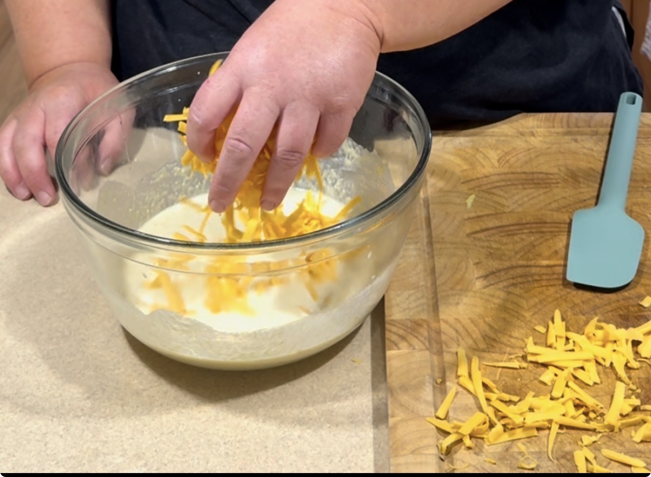 A person’s hand sprinkles shredded cheddar cheese into a glass bowl of milk mixture, with a blue spatula and more shredded cheese on a wooden cutting board nearby.