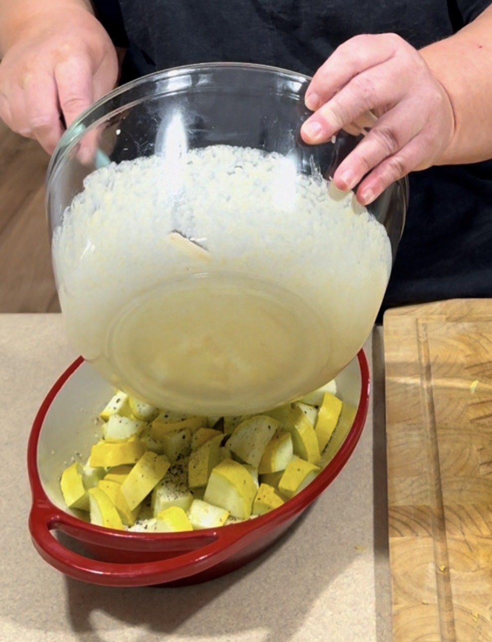 A person pours a creamy mixture from a glass bowl over chopped yellow squash and zucchini in a red baking dish, preparing a casserole on a kitchen counter.