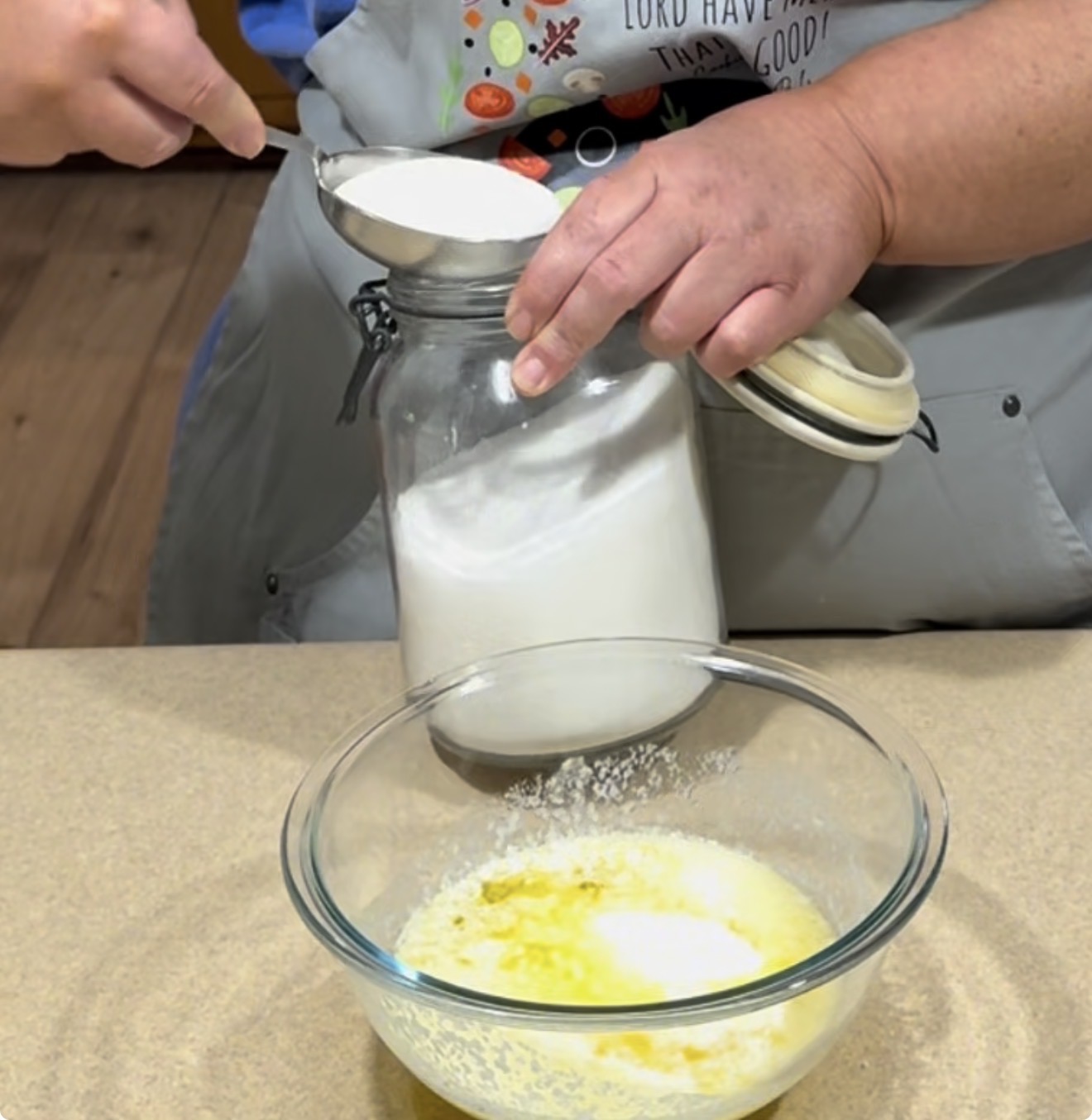 A person scoops sugar from a glass jar into a measuring cup above a mixing bowl containing melted butter on a kitchen counter.