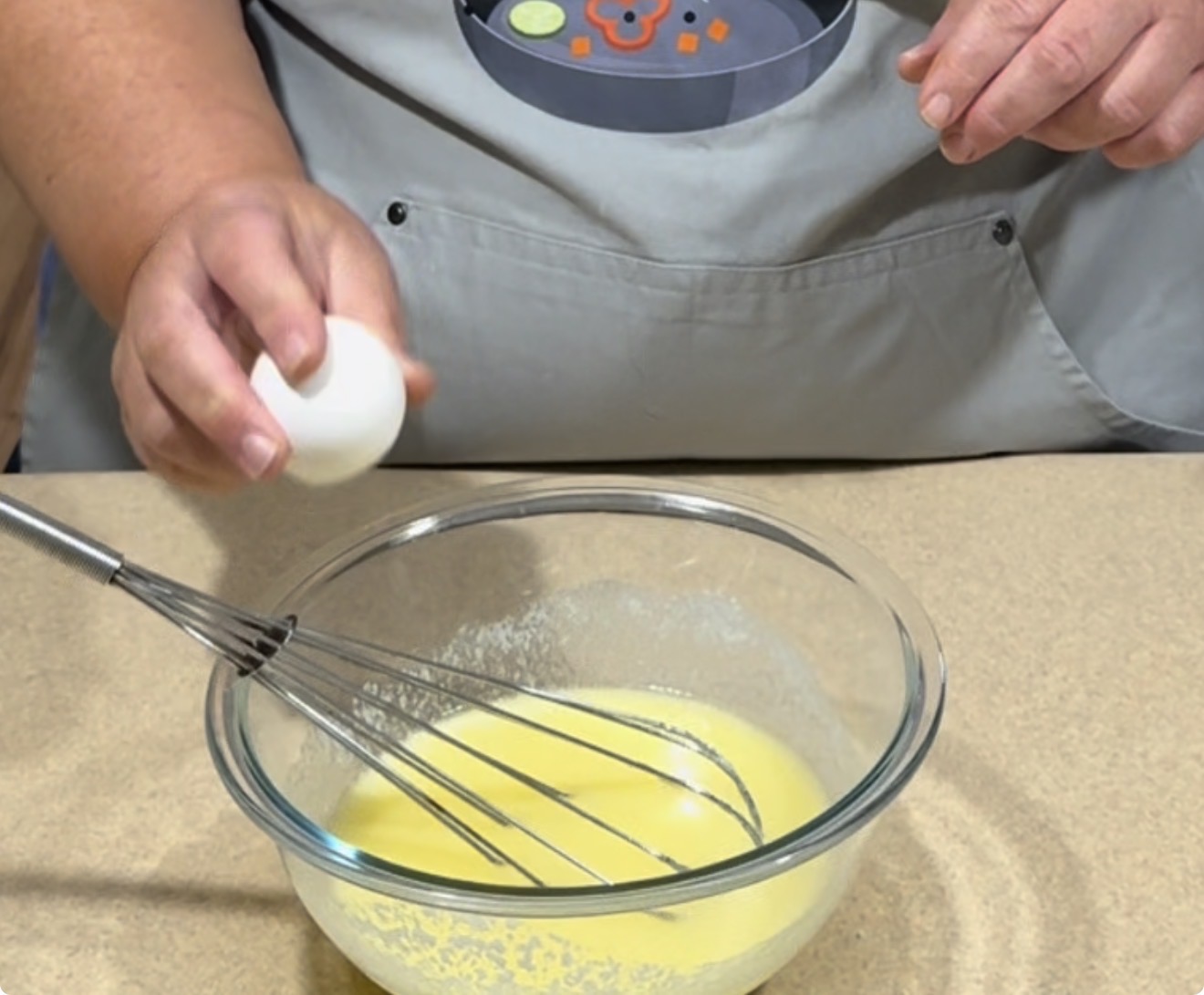 A person wearing a gray apron cracks an egg over a glass bowl of yellow batter, with a whisk resting inside the bowl on a countertop.