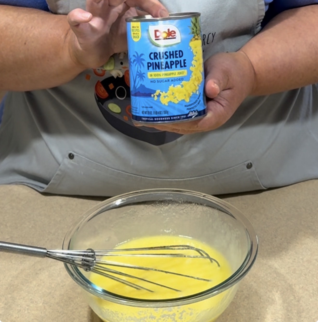 A person in an apron holds a can of Dole crushed pineapple above a glass bowl of yellow mixture. A metal whisk rests in the bowl on a beige countertop.