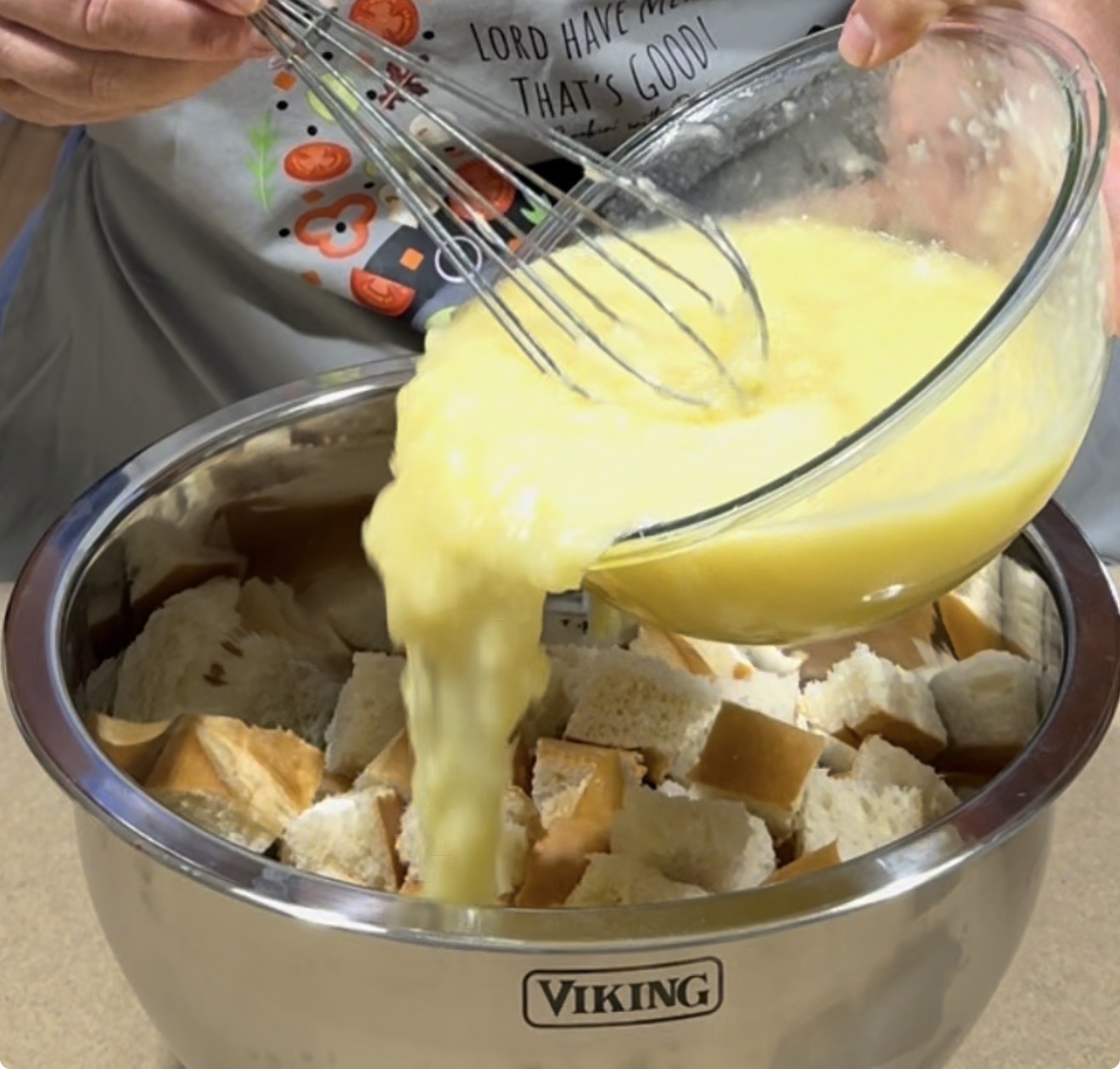 A person pours a bowl of whisked eggs over cubed bread in a large stainless steel mixing bowl labeled Viking, preparing ingredients for a recipe.