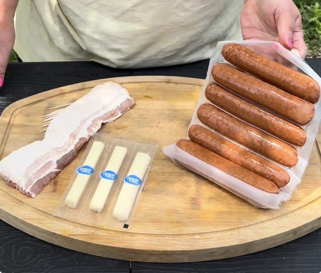 A person holds a pack of six sausages over a wooden cutting board with bacon strips, toothpicks, and two wrapped mozzarella cheese sticks placed on it.