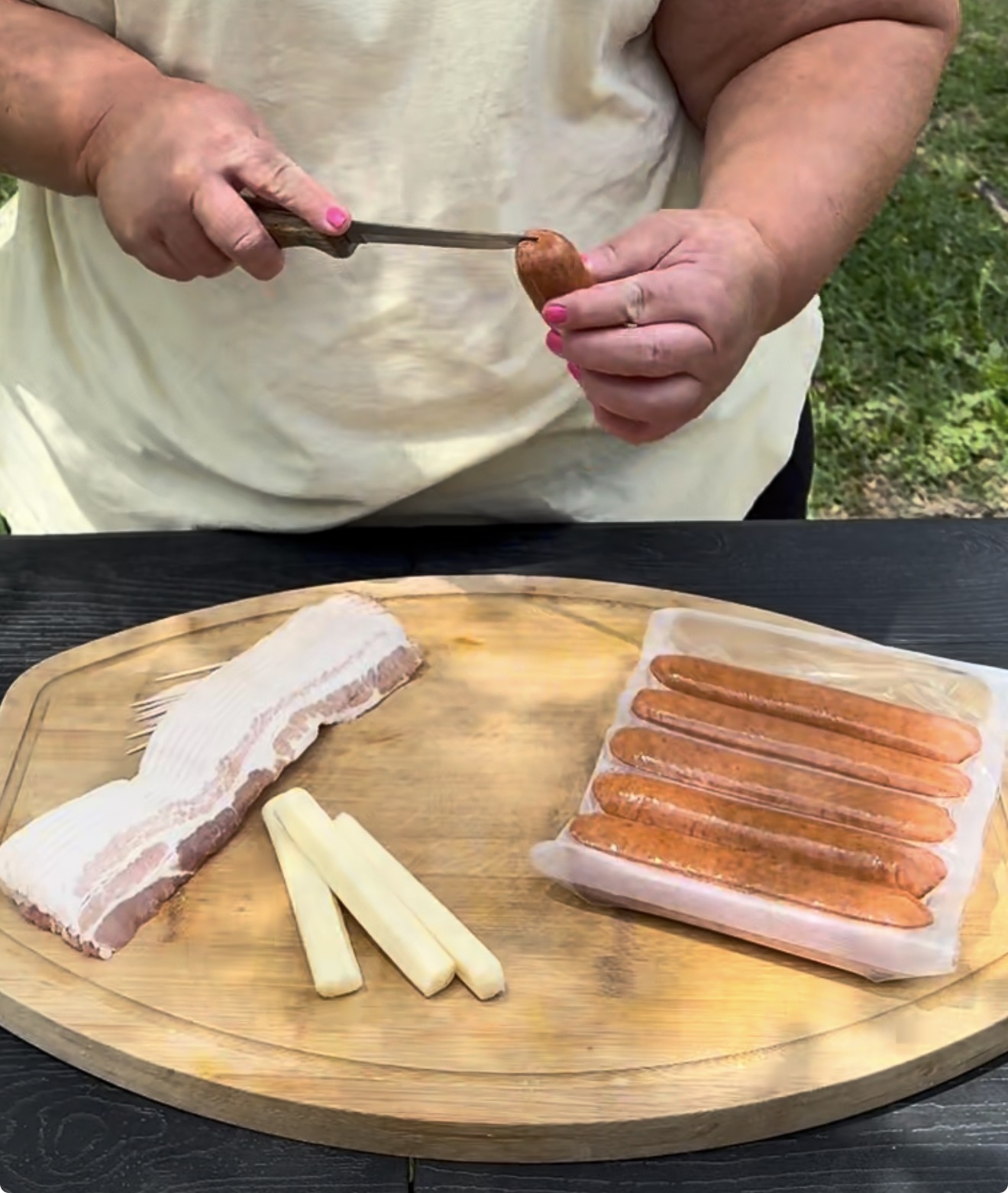 A person slices a sausage with a knife. On a wooden board are strips of bacon, cheese sticks, and a package of sausages, outdoors on a sunny day.