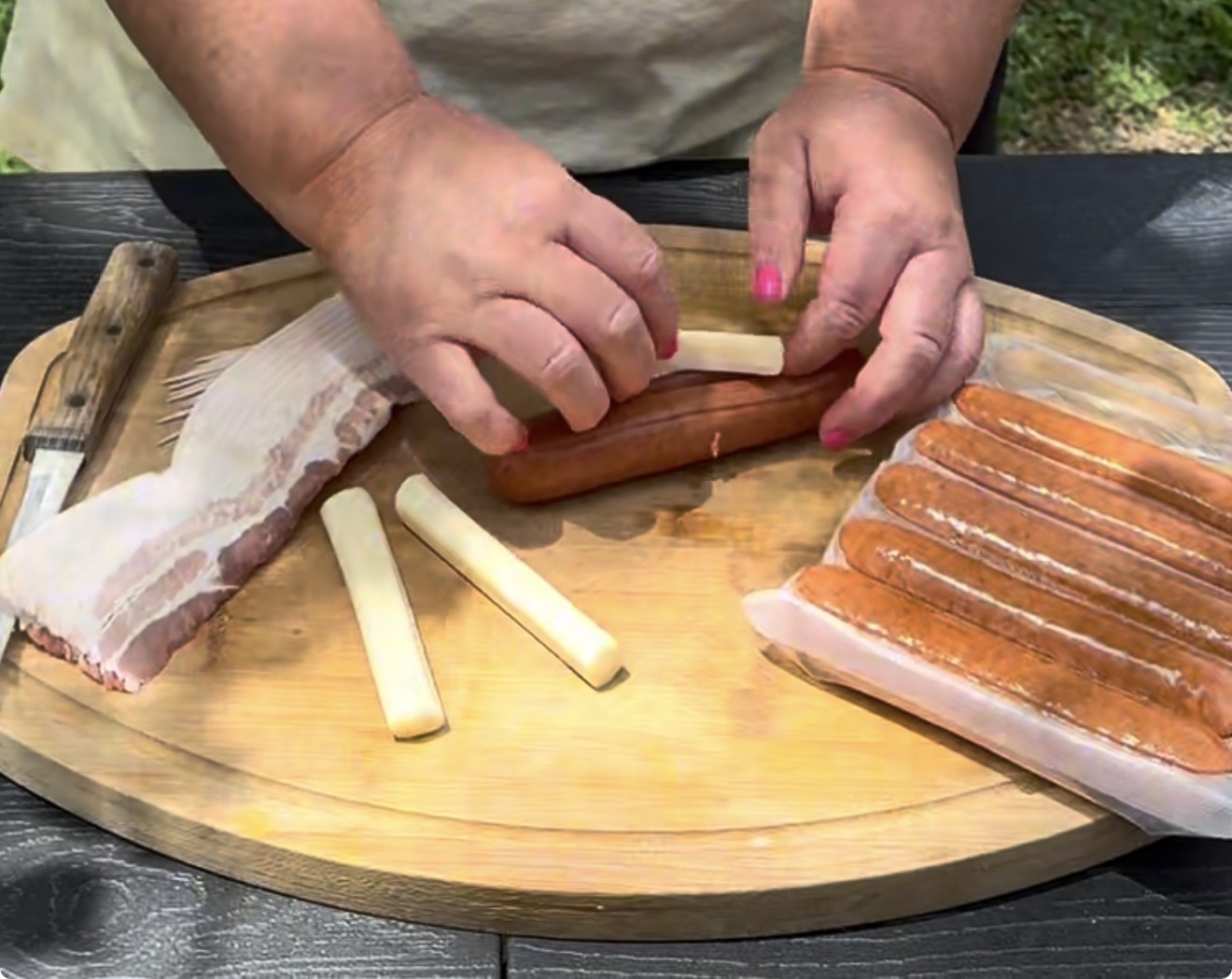A person prepares food outdoors, stuffing a hot dog with a piece of cheese on a wooden cutting board. Nearby are hot dogs, bacon strips, and a knife.