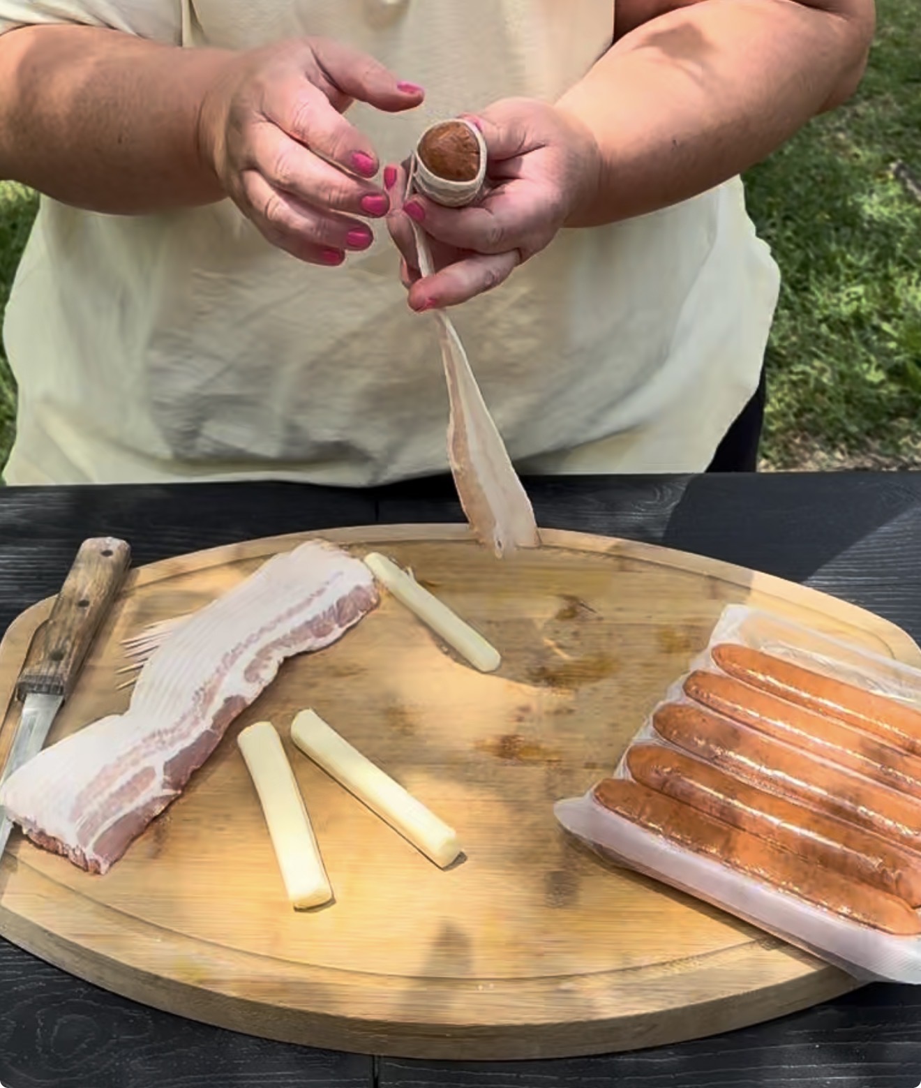 A person wraps a hot dog with a slice of bacon at an outdoor table. On the wooden cutting board are bacon strips, cheese sticks, and hot dogs. Grass is visible in the background.