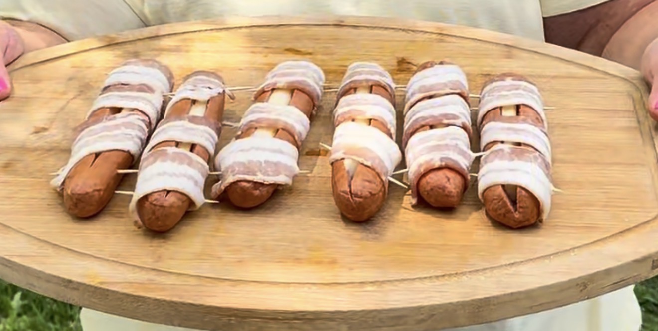 A person holds a wooden platter with six hot dogs, each wrapped with several strips of uncooked bacon. The hot dogs have slits cut into them and are arranged neatly in two rows.