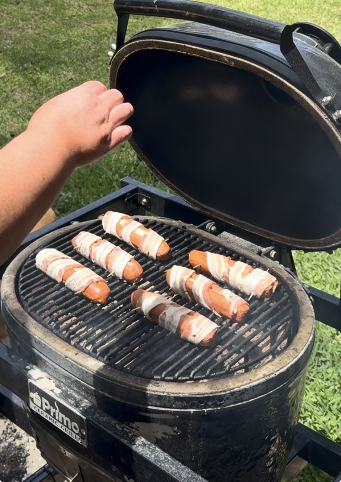 A hand lifts the lid of a black Primo grill where seven bacon-wrapped hot dogs are cooking on the grate. Smoke rises as sunlight shines on the grass in the background.