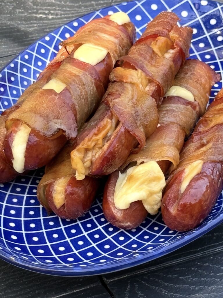 Four grilled sausages stuffed with melted cheese and wrapped in crispy bacon are arranged on a blue, patterned plate. The cheese is oozing out of the sausages in some spots.