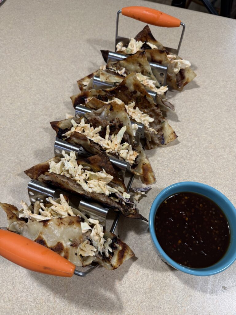 A row of crispy tacos filled with shredded meat and cheese sits in metal taco holders on a countertop, with a small blue bowl of dark dipping sauce beside them.