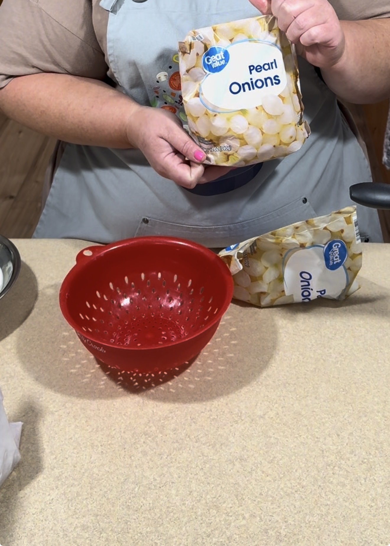 A person holding a bag of Great Value pearl onions above a countertop, next to a red colander and another bag of pearl onions. The person is wearing an apron.