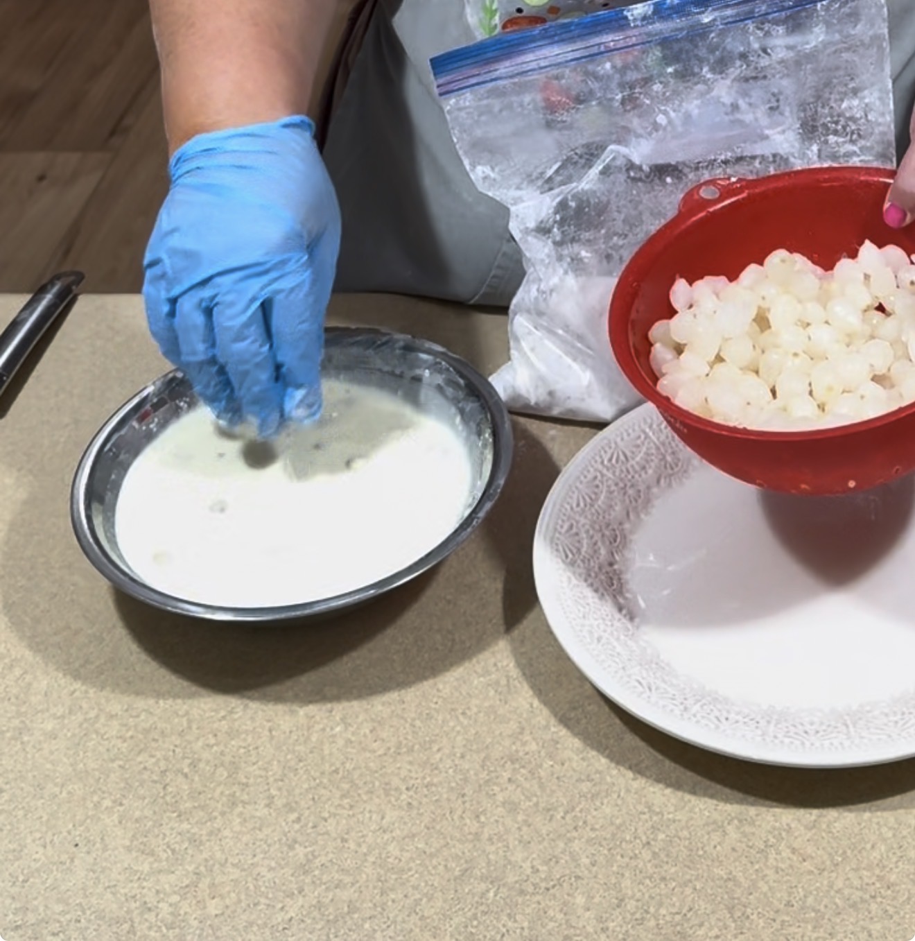 A person wearing a blue glove dips a hand into a bowl of white liquid next to a red bowl filled with white, round pieces on a kitchen counter. A large bag and an empty plate are also visible.