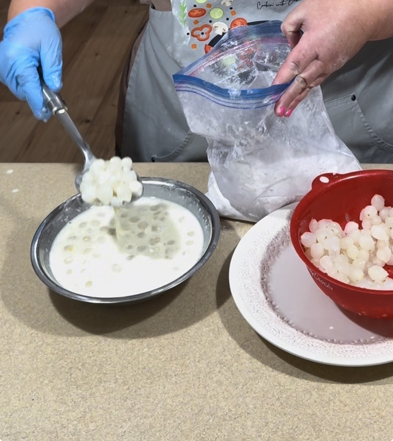 A person transfers small white tapioca pearls from a metal bowl of milky liquid into a red strainer, with a bag of flour and more tapioca pearls on the counter.