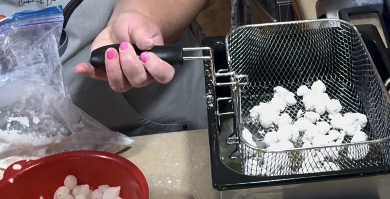 A person with pink-painted nails lowers a fryer basket filled with battered pieces of food into a deep fryer. A red bowl and a bag with similar food pieces are on the counter nearby.