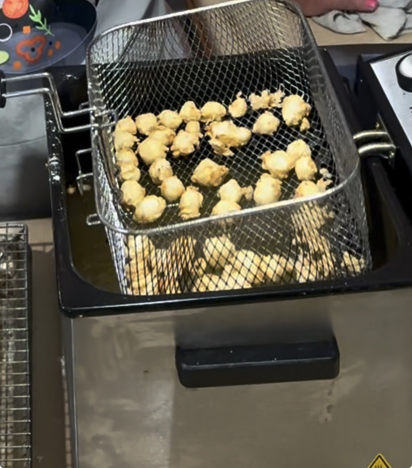 A metal basket filled with small, golden-brown battered food pieces is being lifted from a deep fryer, with hot oil visible beneath the basket.