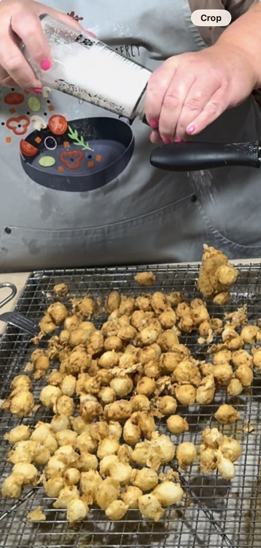 A person with pink nail polish sprinkles salt from a grinder onto fried, golden-brown cauliflower bites on a cooling rack. A gray apron with food graphics is visible in the background.