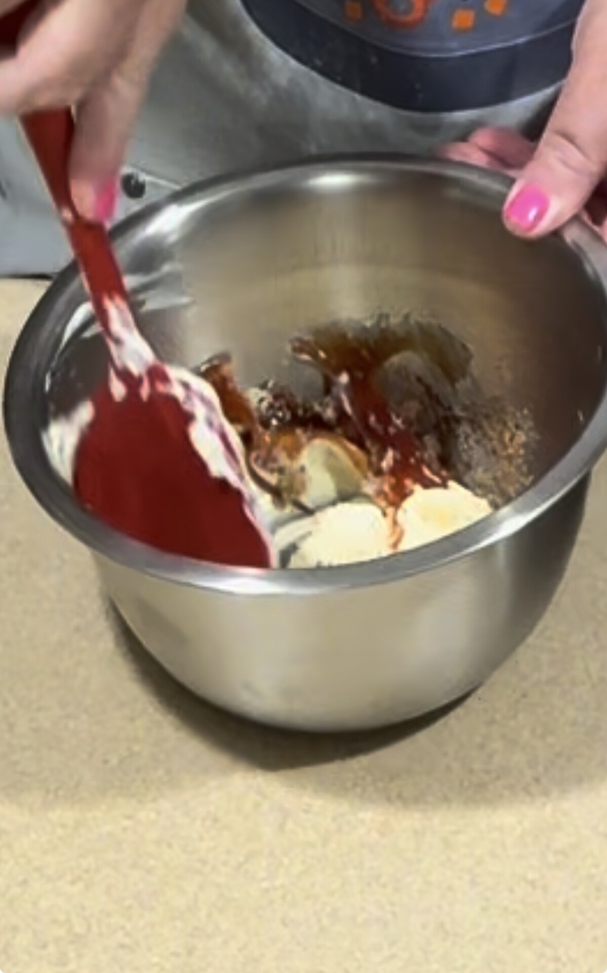 A person with pink nail polish uses a red spatula to mix ingredients, including brown sugar and vanilla, in a stainless steel bowl on a countertop.