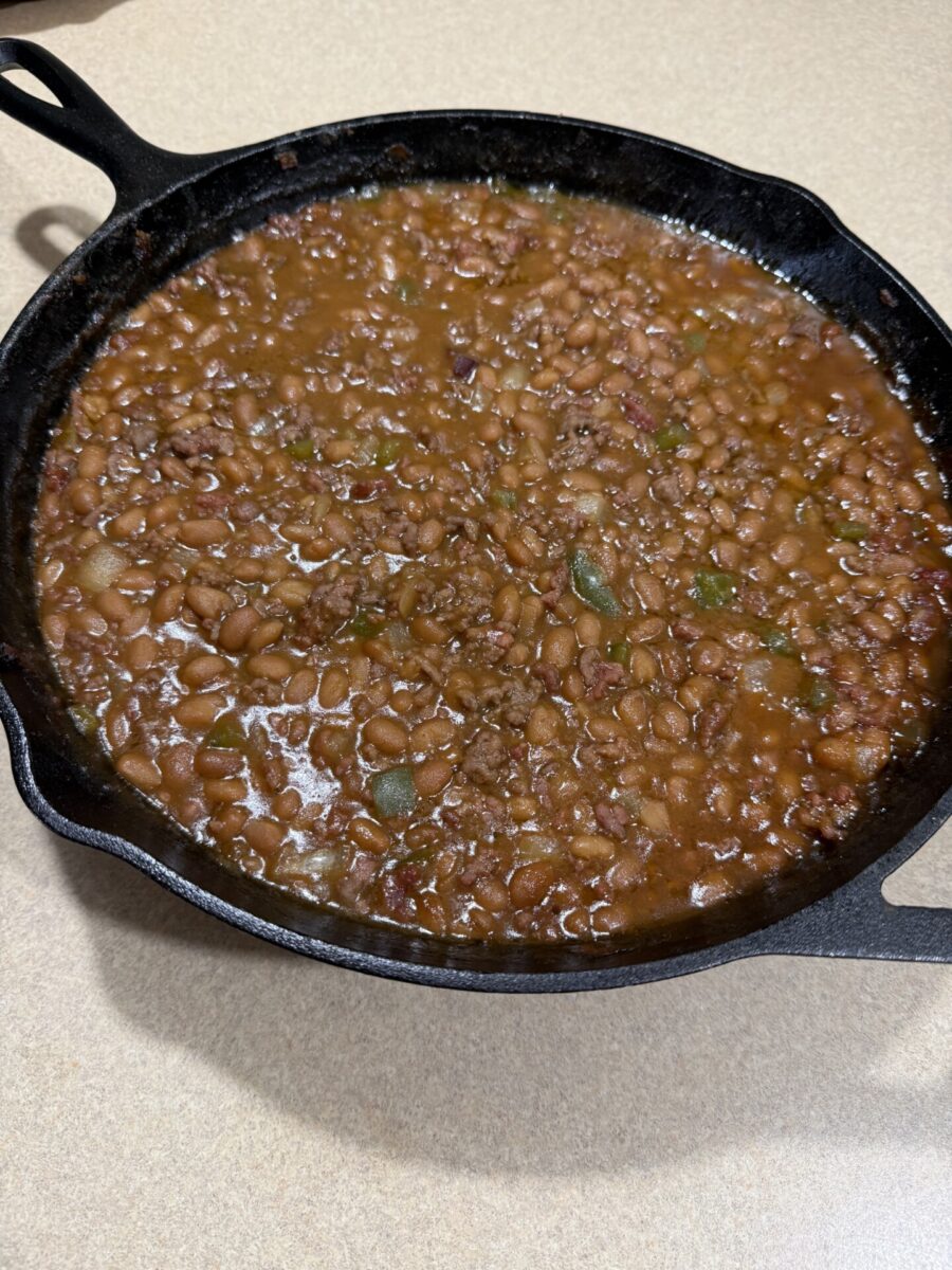 A cast iron skillet filled with cooked beans in a thick, brown sauce, mixed with pieces of green pepper and ground meat, sitting on a beige countertop.