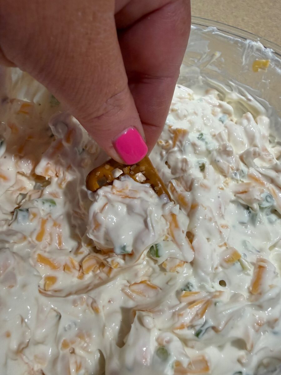A hand with pink nail polish dips a pretzel into a creamy mixture with shredded cheese and green herbs in a glass bowl.