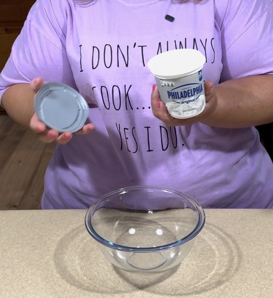 A person wearing a lavender shirt that says “I DON’T ALWAYS COOK… YES I DO!” holds an open container of Philadelphia cream cheese above a clear glass mixing bowl on a kitchen counter.