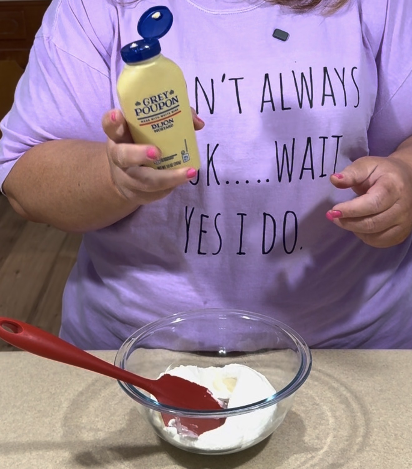 A person wearing a lavender shirt with text holds a bottle of Grey Poupon Dijon mustard above a mixing bowl with a red spatula and white ingredients on a kitchen counter.